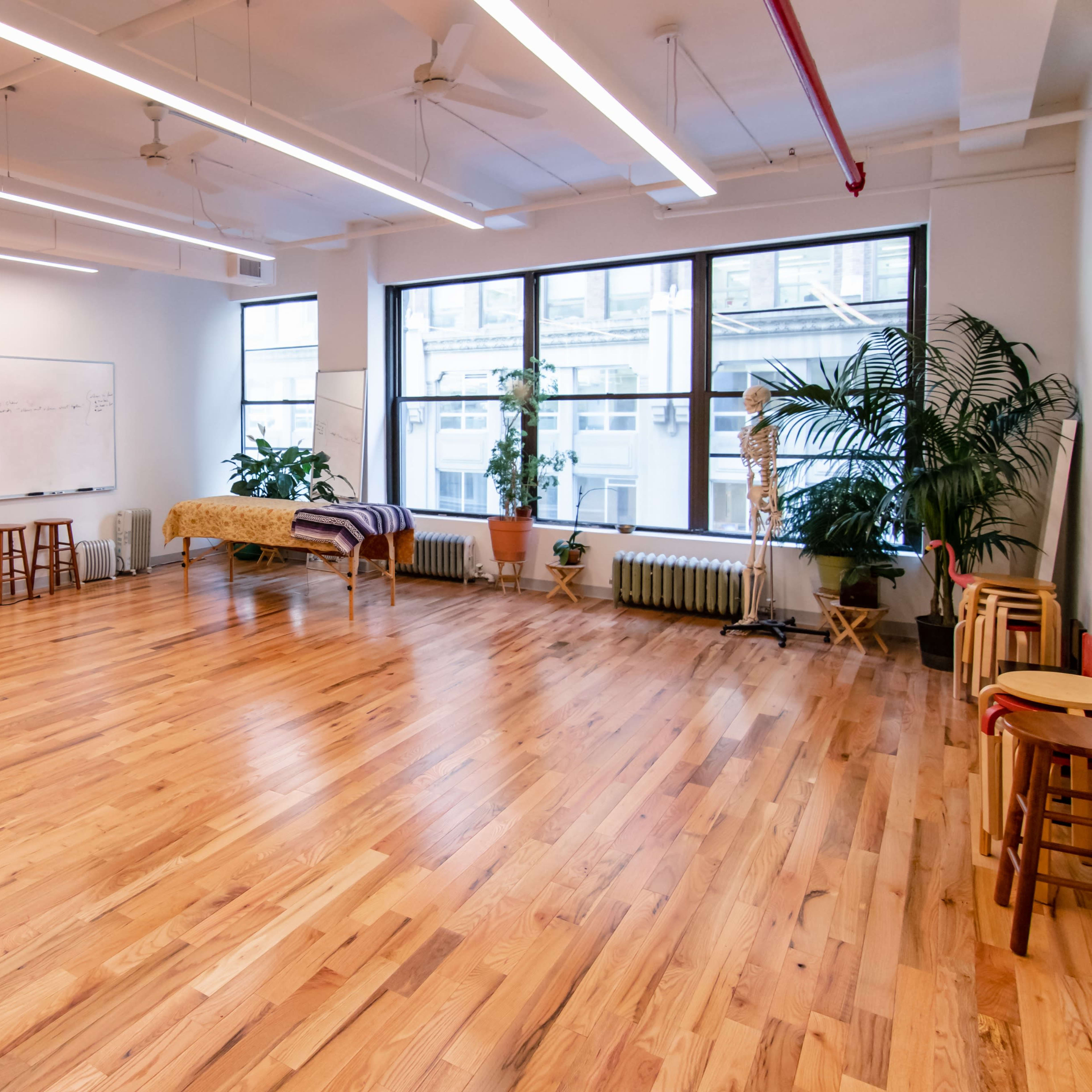 A spacious studio with wooden flooring, large windows, several chairs, and a whiteboard on the wall.