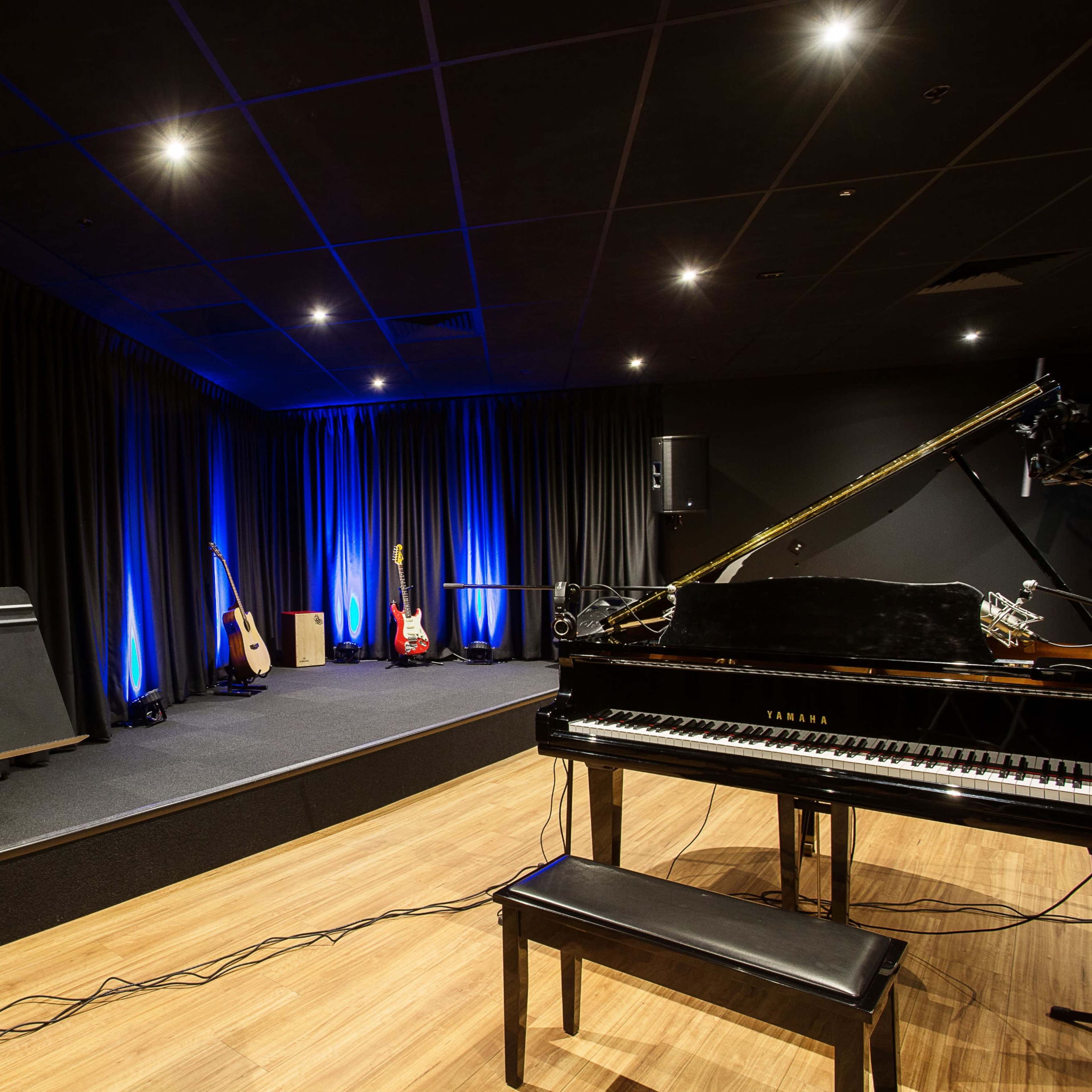 A music studio featuring a grand piano, microphones, audio equipment, and illuminated blue lights against a black curtain backdrop.