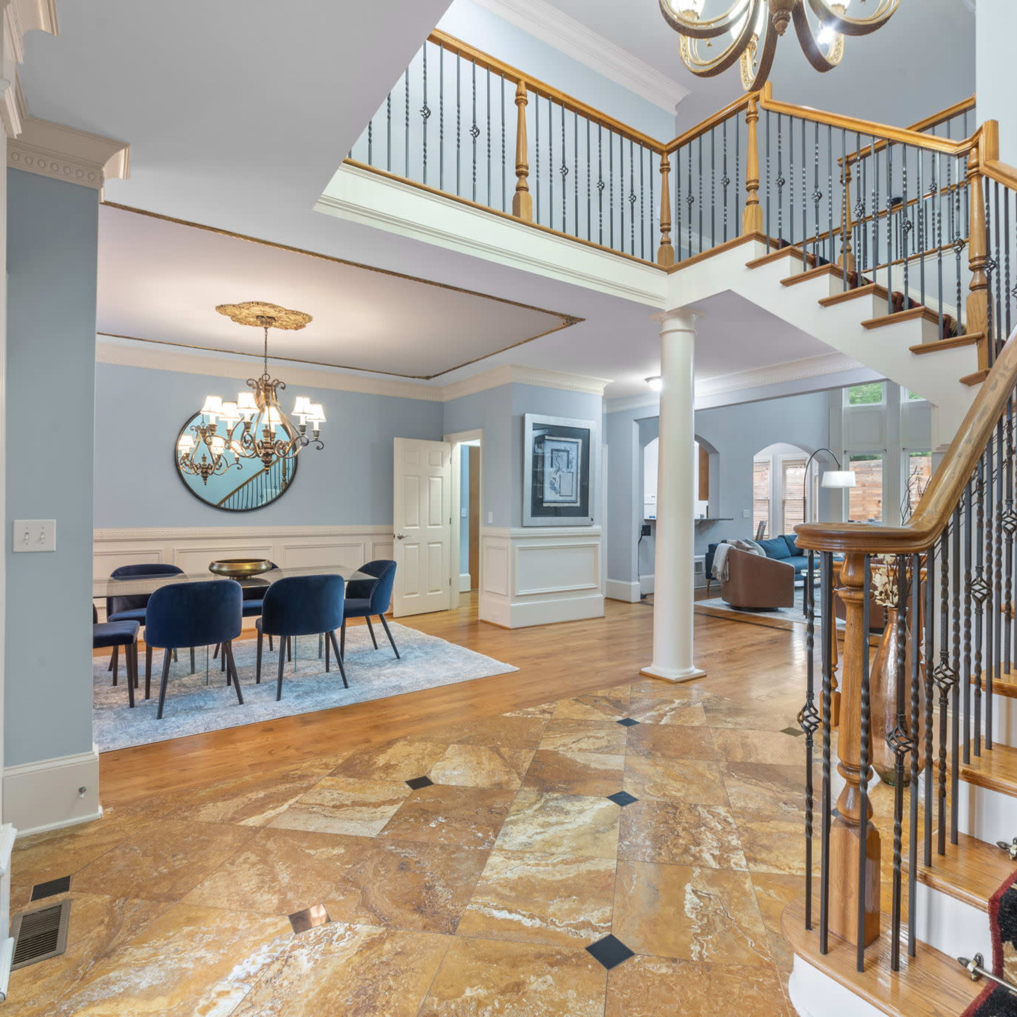 A spacious foyer with a winding staircase, hardwood flooring, and a chandelier, leading to a dining area in the background.