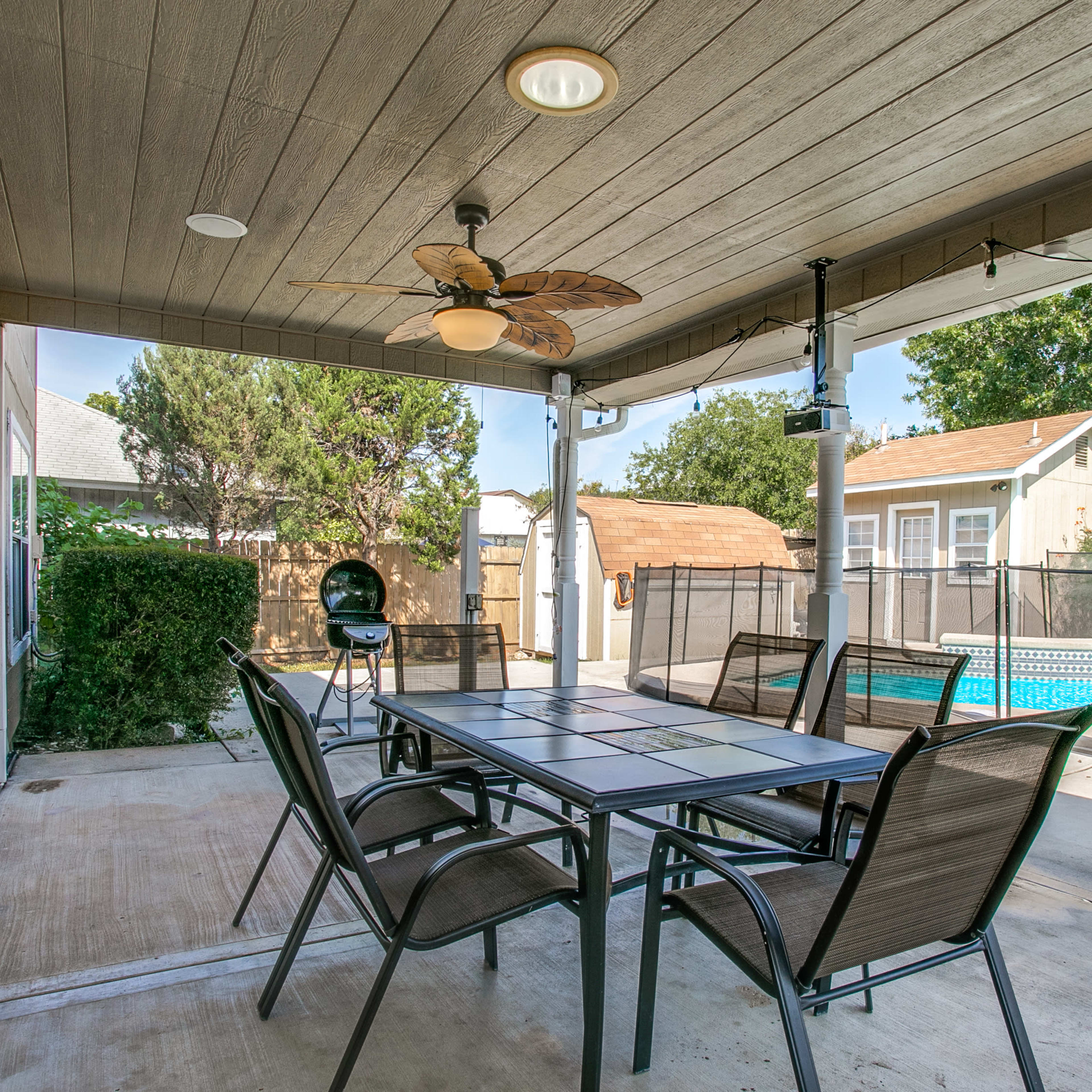 A covered outdoor patio with a dining table, chairs, a ceiling fan, and a view of a swimming pool surrounded by a fence.