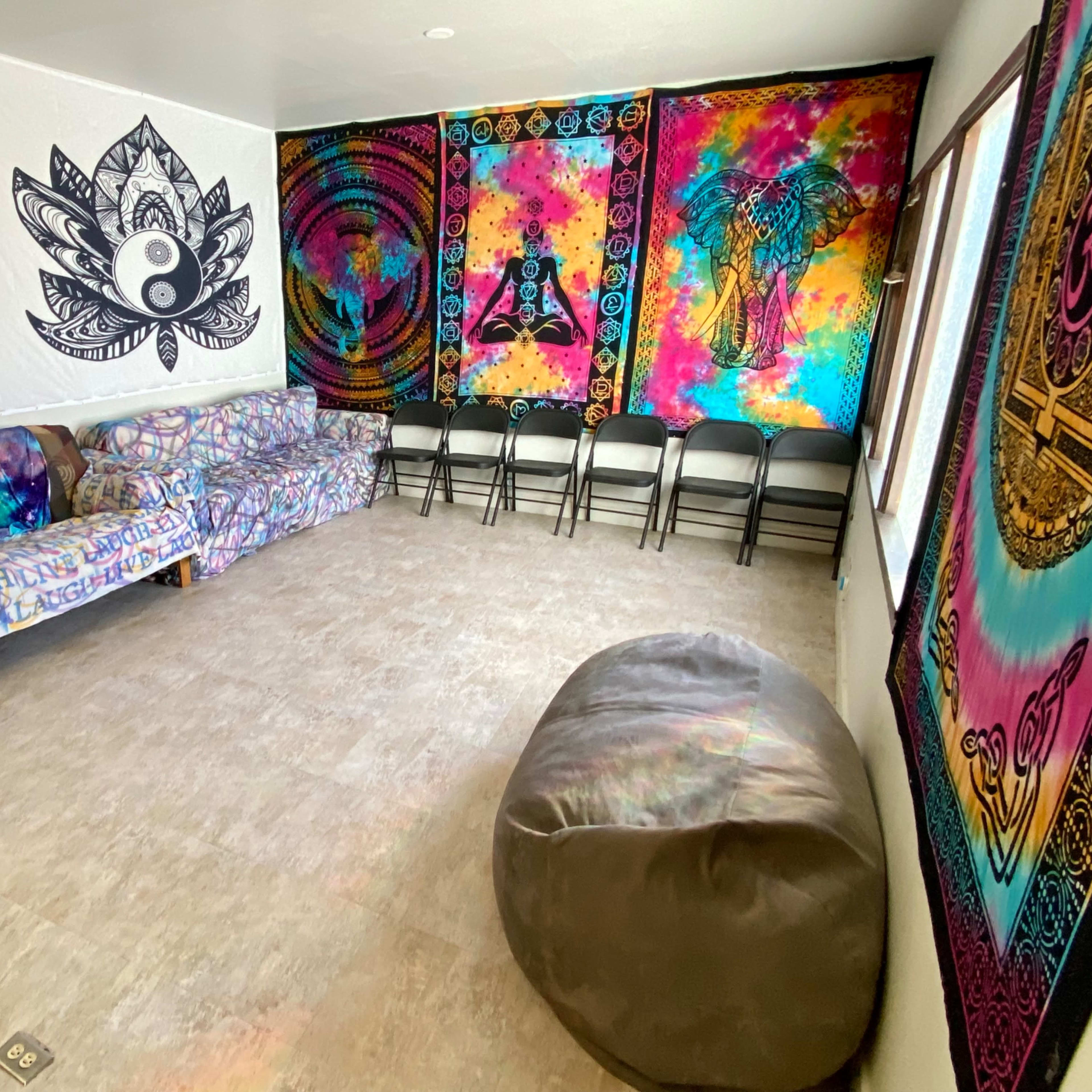A brightly colored room with patterned wall tapestries, a sofa covered in a printed throw, several folding chairs, and a large bean bag on the floor.