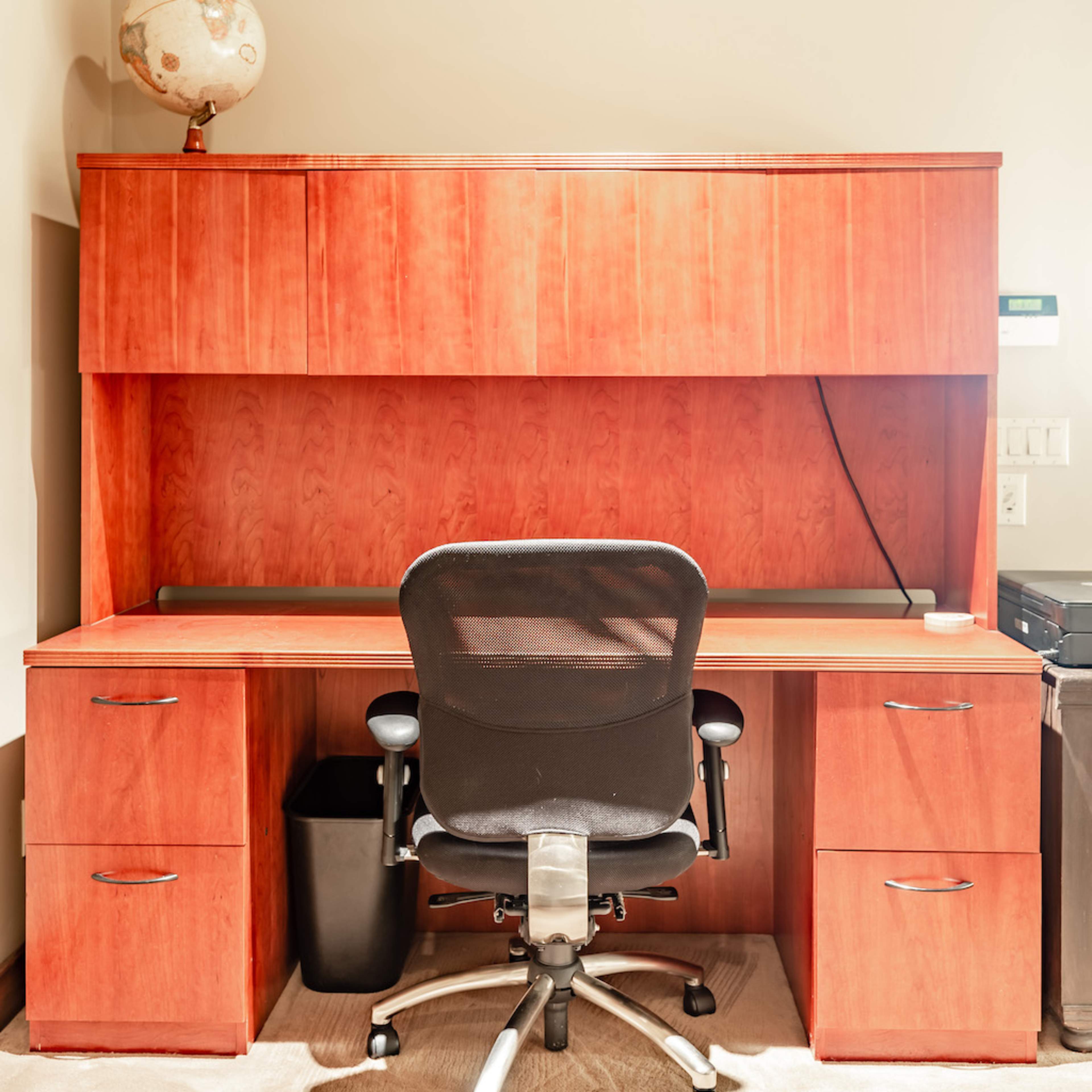 An empty office desk setup with a black chair, wooden cabinetry, and a globe on top, positioned against a wall.