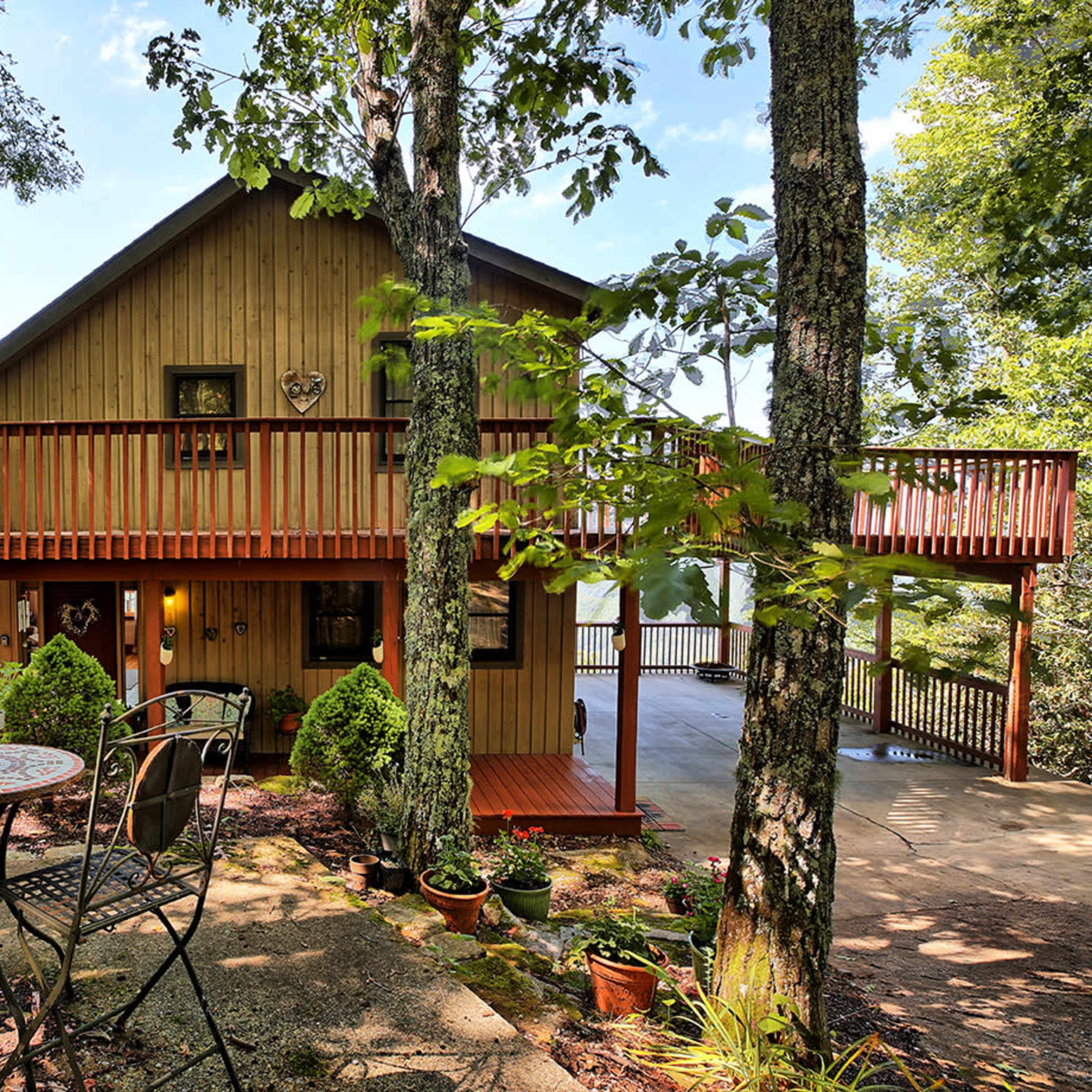 A wooden house with a balcony is surrounded by trees and features a small patio with a table and chairs in front.