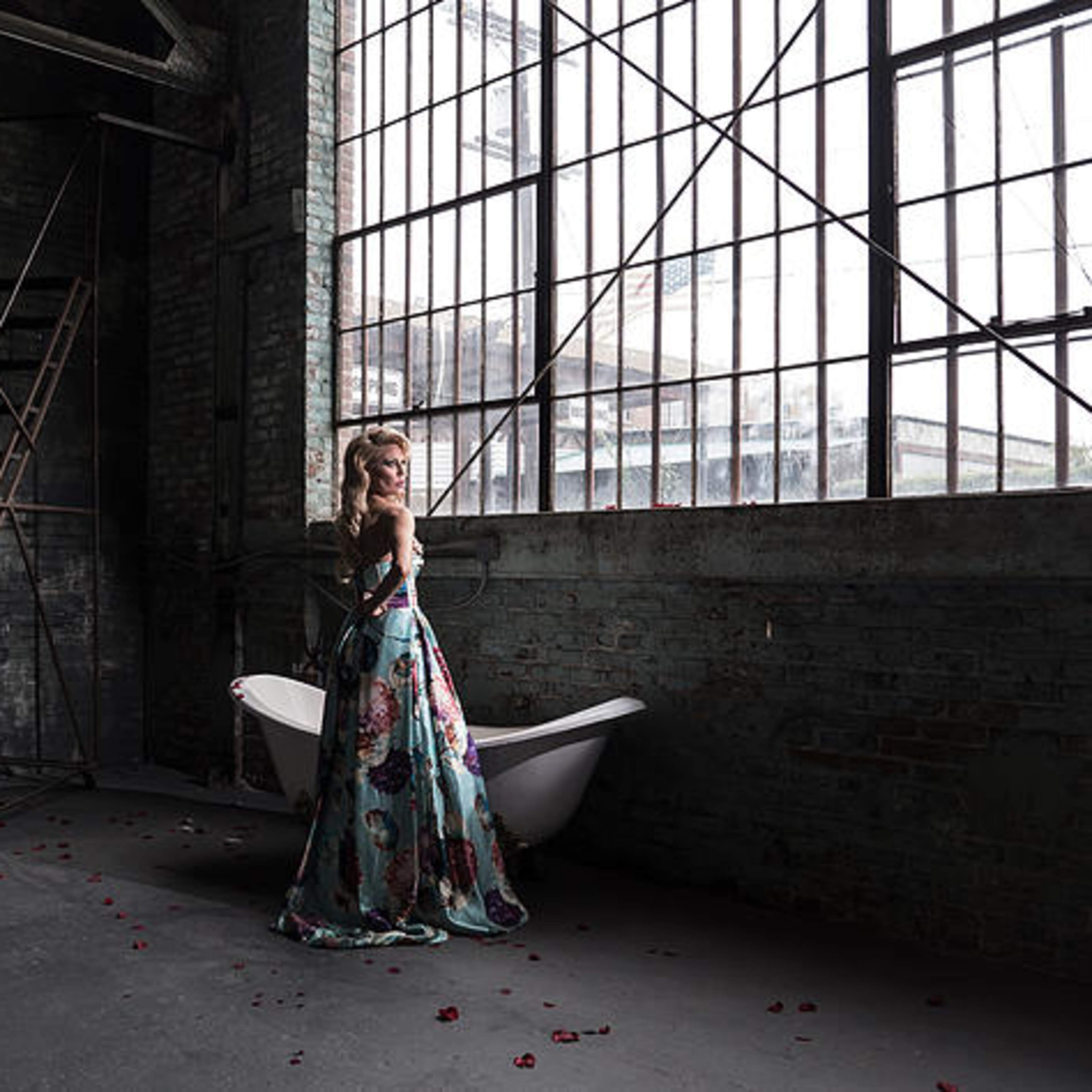A woman in a floral gown stands beside a vintage bathtub in a large, industrial room with tall windows and a metal ladder.