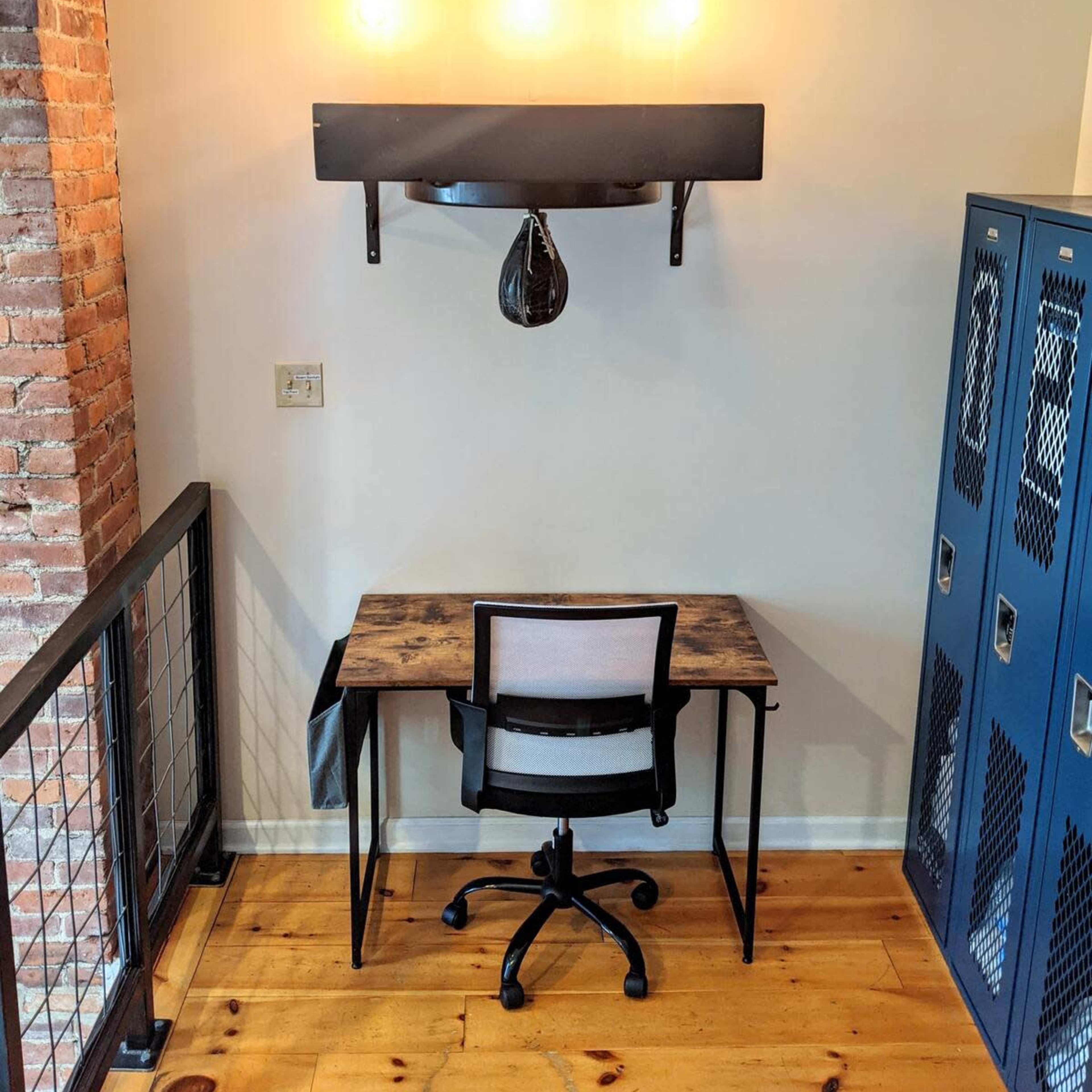 A workspace featuring a small desk and chair beneath a shelf with hanging light bulbs and a boxing speed bag, next to blue lockers and a brick wall.