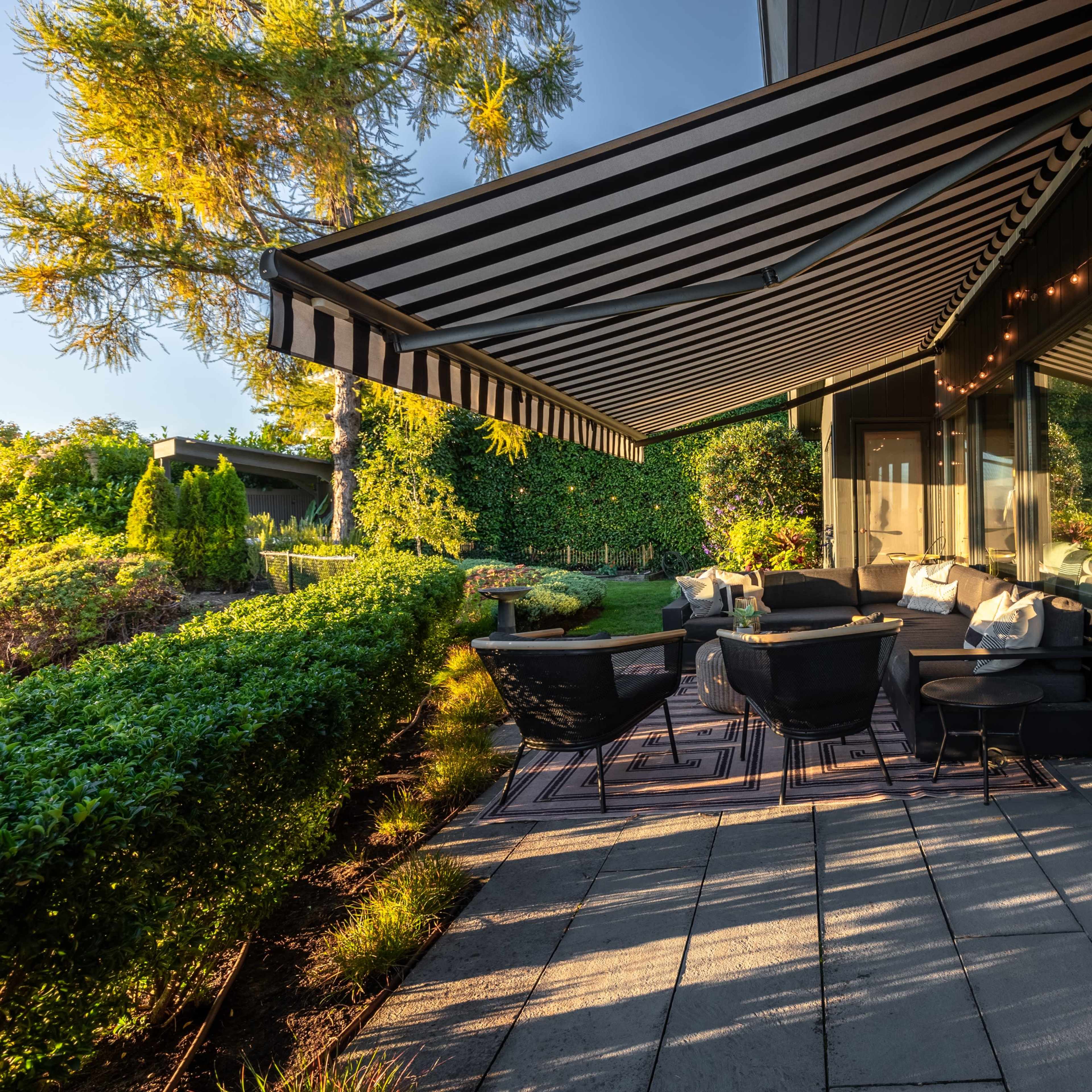 A patio with a striped awning is furnished with wicker chairs and surrounded by neatly trimmed greenery.