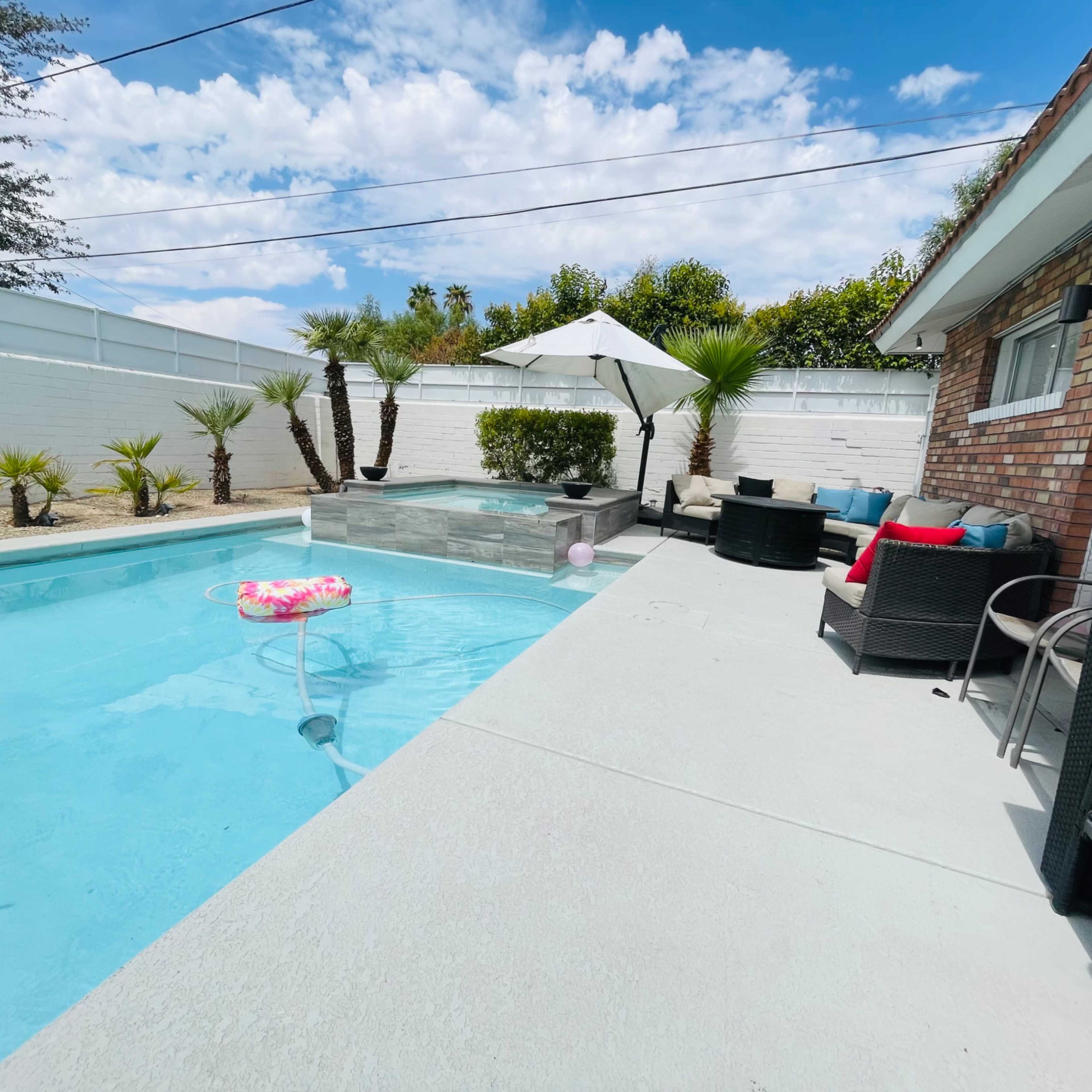 A residential backyard featuring a swimming pool, a spa, palm trees, and outdoor seating with colorful cushions.