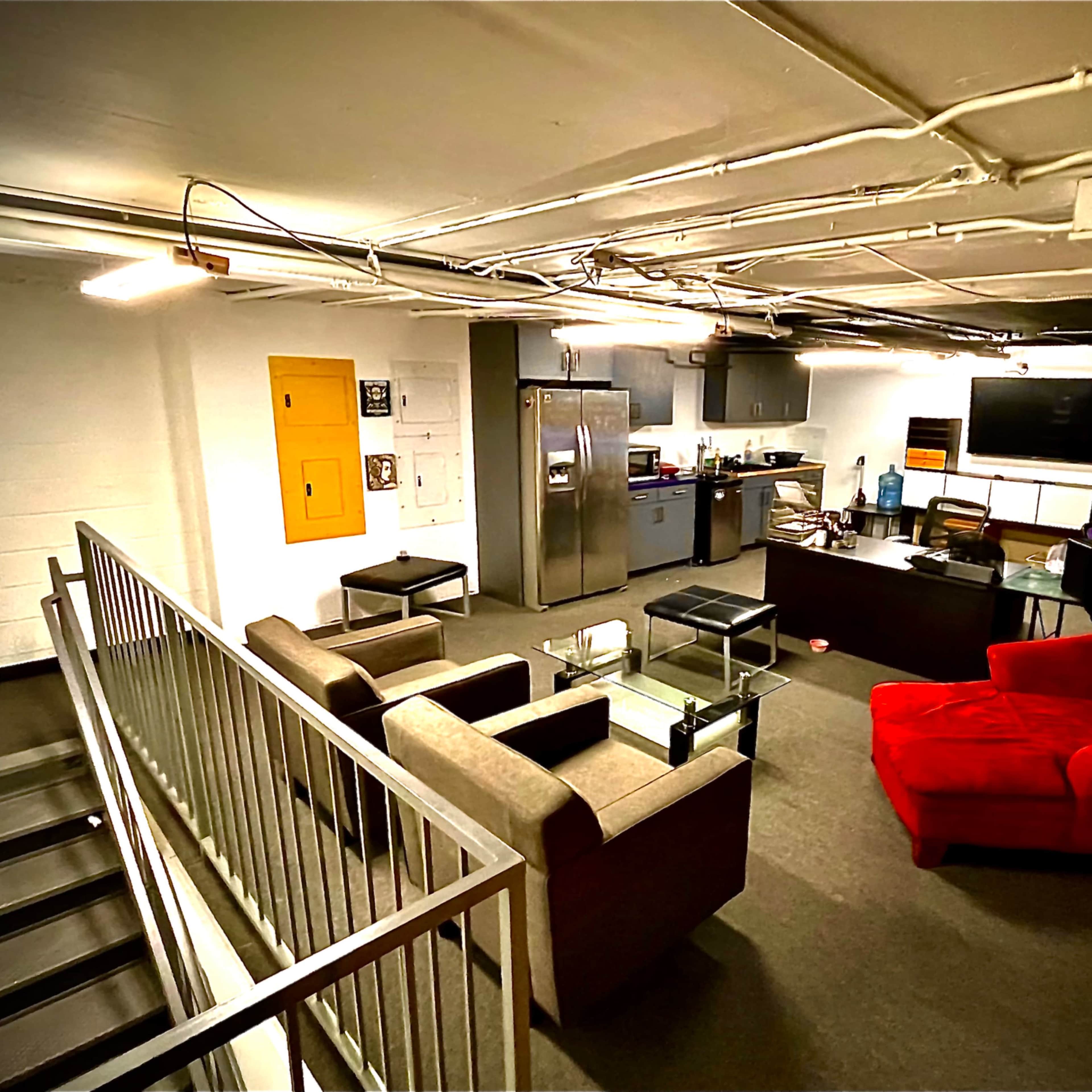 A basement area furnished with a red couch, gray chairs, a kitchen space with stainless steel appliances, and a desk with a computer monitor.