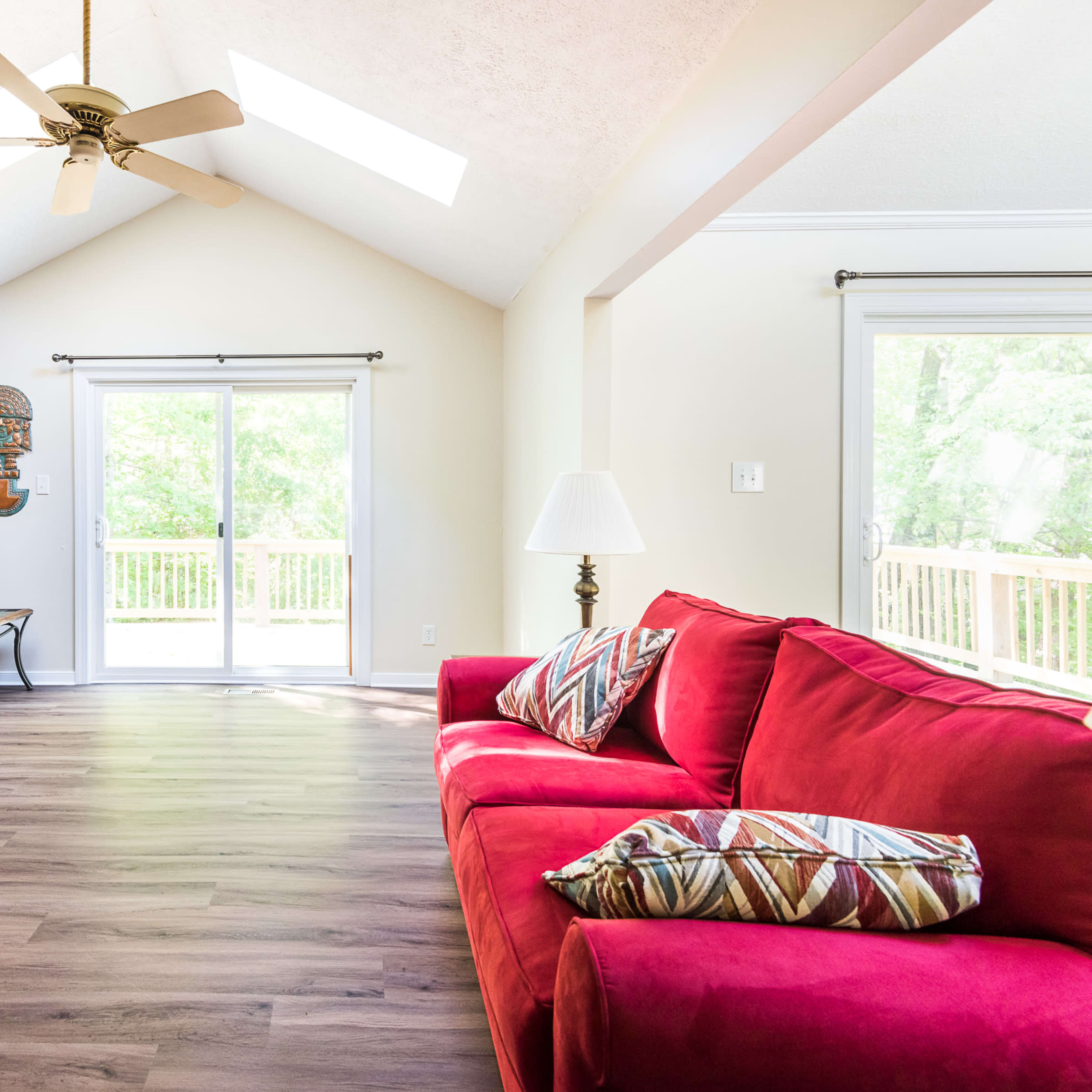 A living room featuring a red sofa with patterned pillows, a wooden accent wall, and large windows leading to a deck outside.