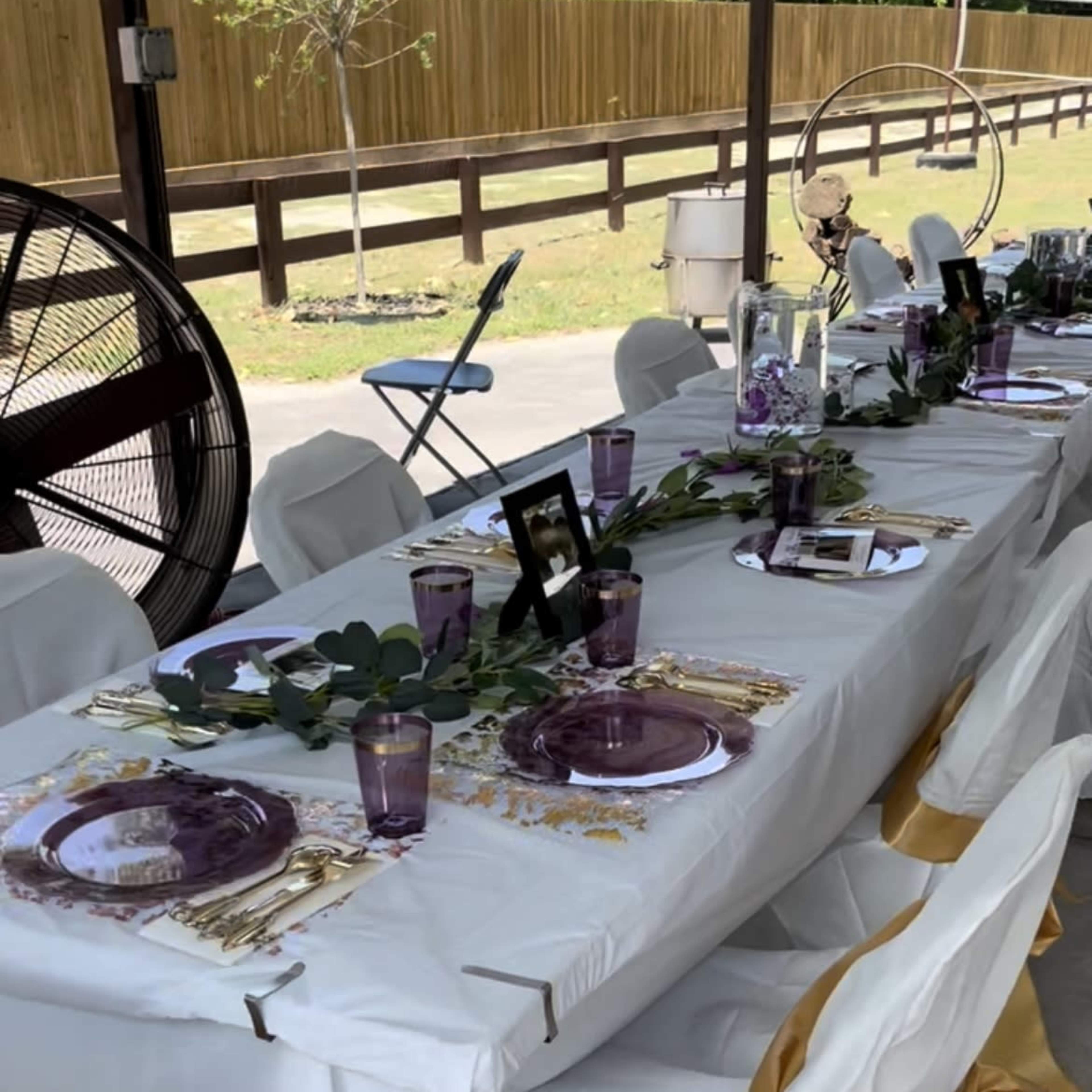 A long table is set for a gathering, featuring white tablecloths, purple decorations, and floral arrangements under a covered outdoor area.