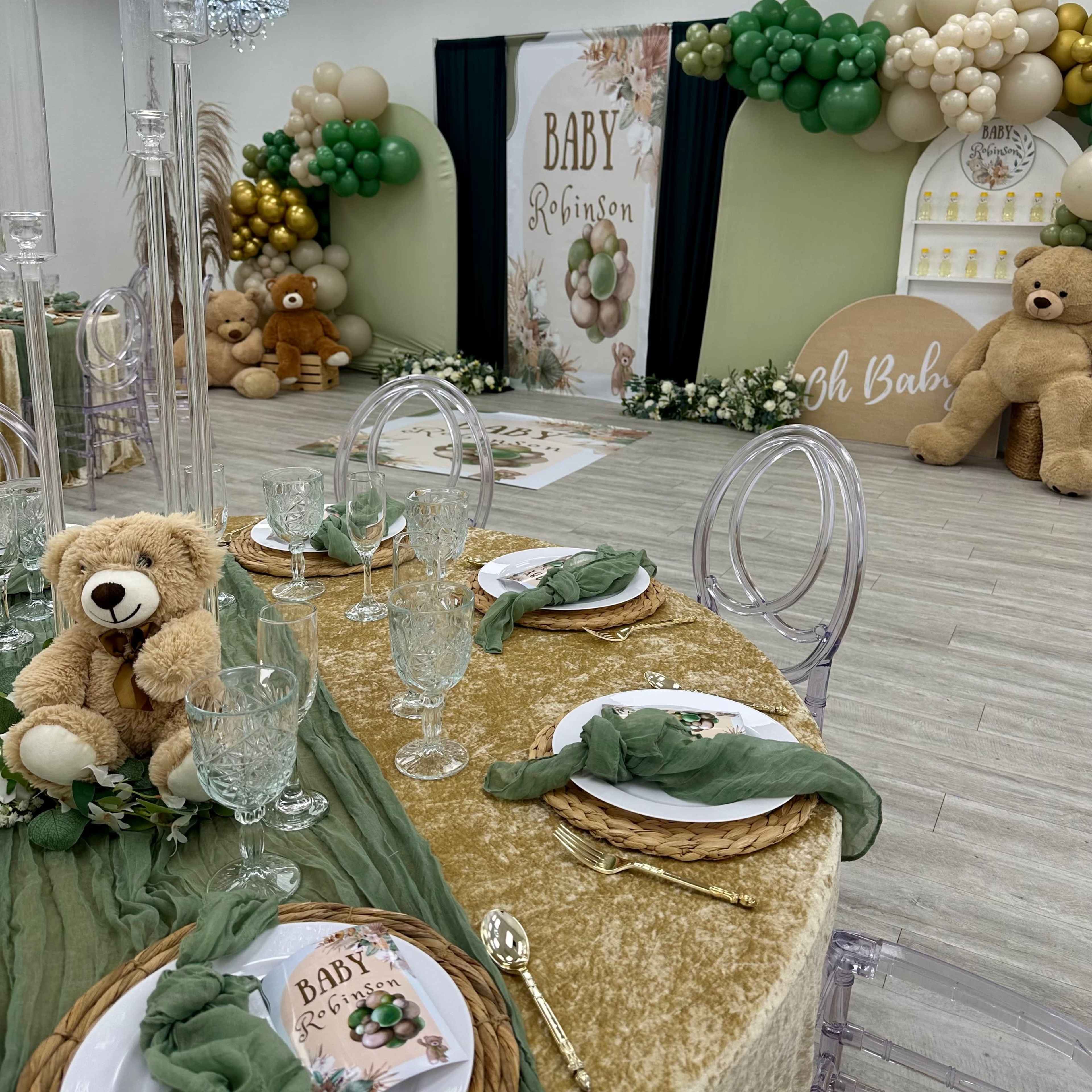 A decorated event space features a table set with elegant glassware and greenery, alongside a backdrop with a "Baby Robinson" theme, complemented by balloons and plush teddy bears.