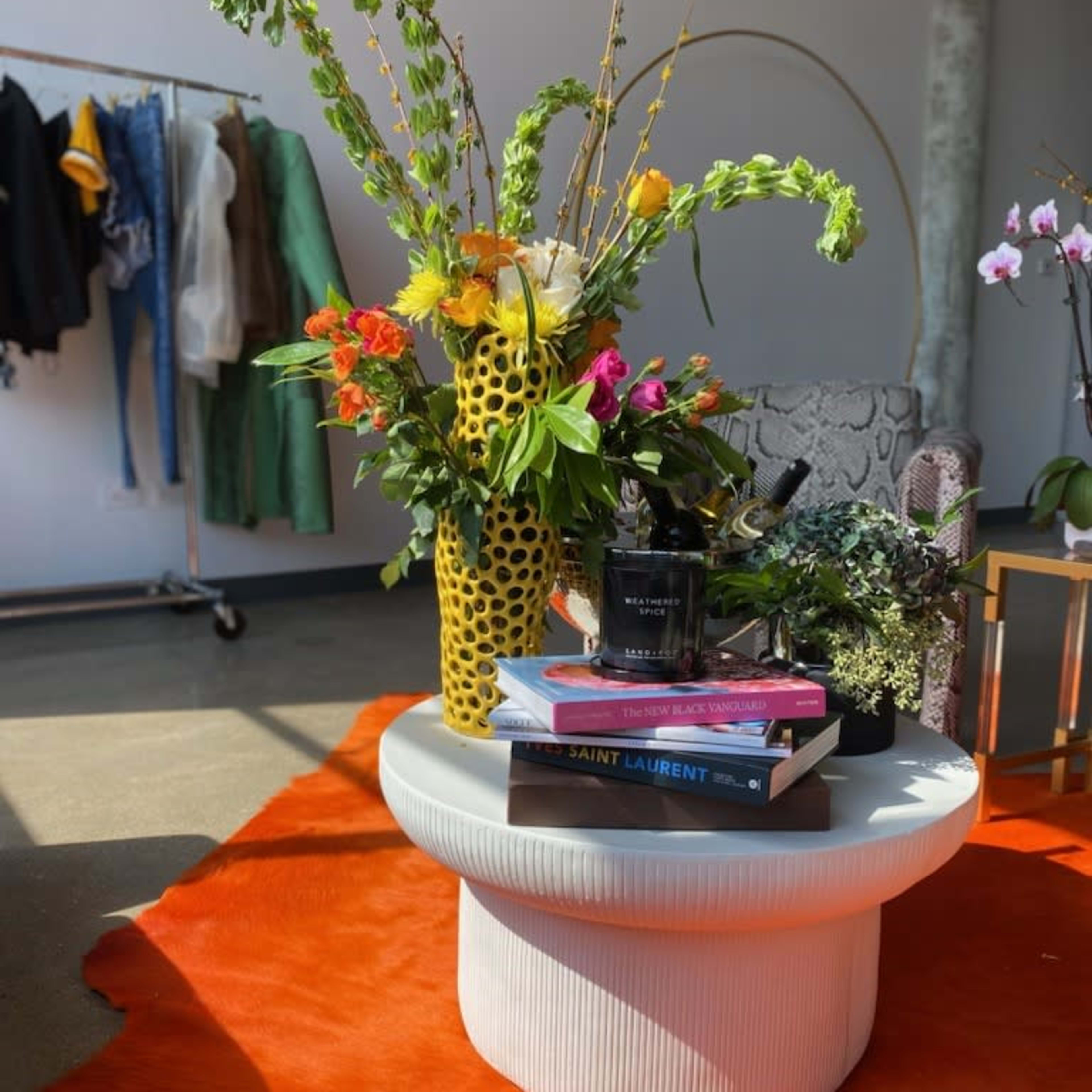 A round coffee table topped with books, a candle, and a vibrant floral arrangement sits on a bright orange rug in a styled room with clothing on a rack in the background.