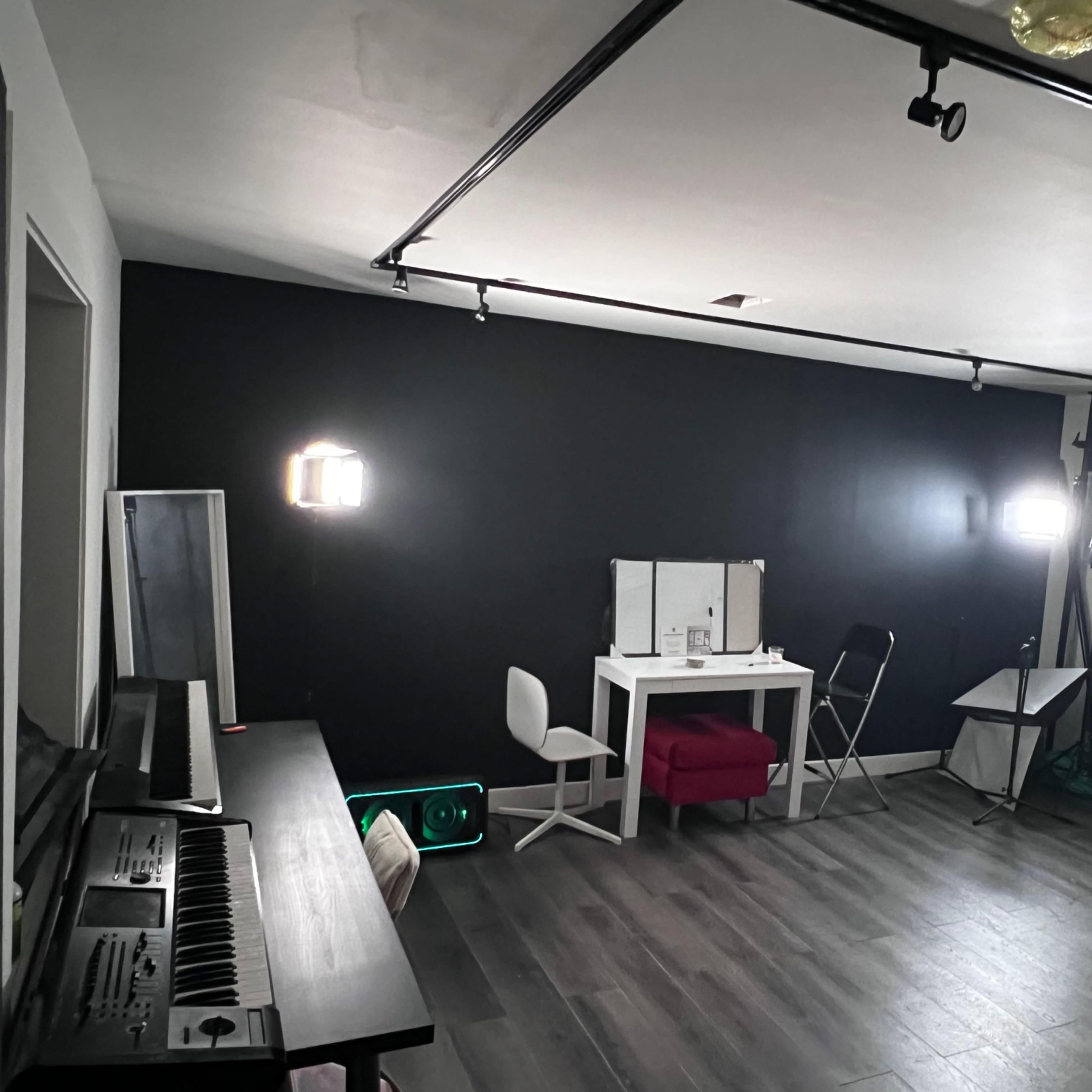 A room with a black accent wall features a white table, chairs, a piano, and studio lights set up for filming or photography.