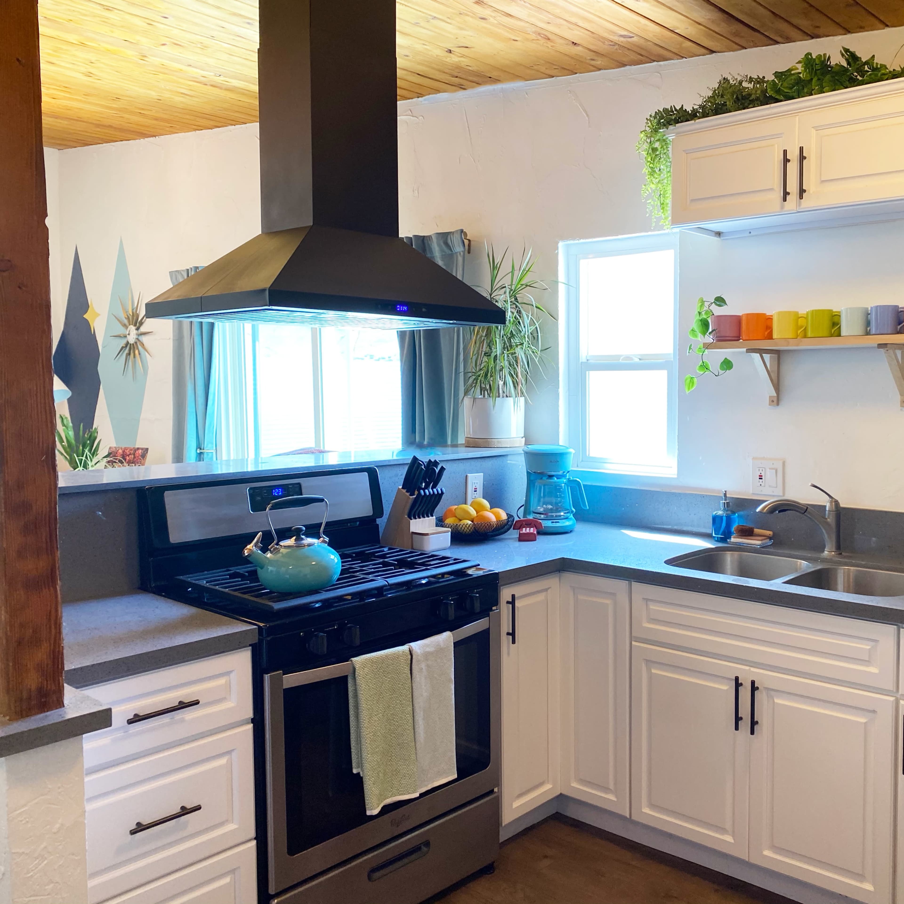 A modern kitchen features a stainless steel stove, a silver refrigerator, and white cabinetry, with a window allowing natural light and colorful dishware displayed on a shelf.
