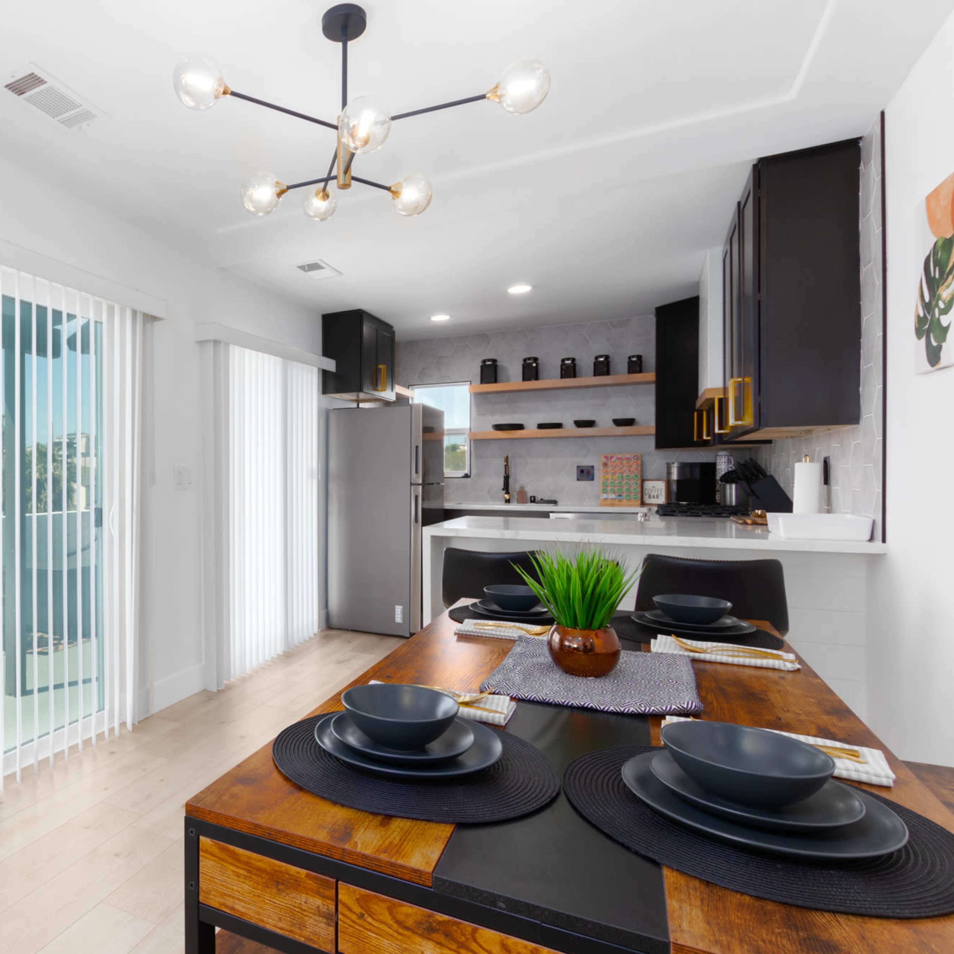 A modern kitchen and dining area feature a wooden table set for four, with a stylish light fixture above and large glass doors leading to a balcony.