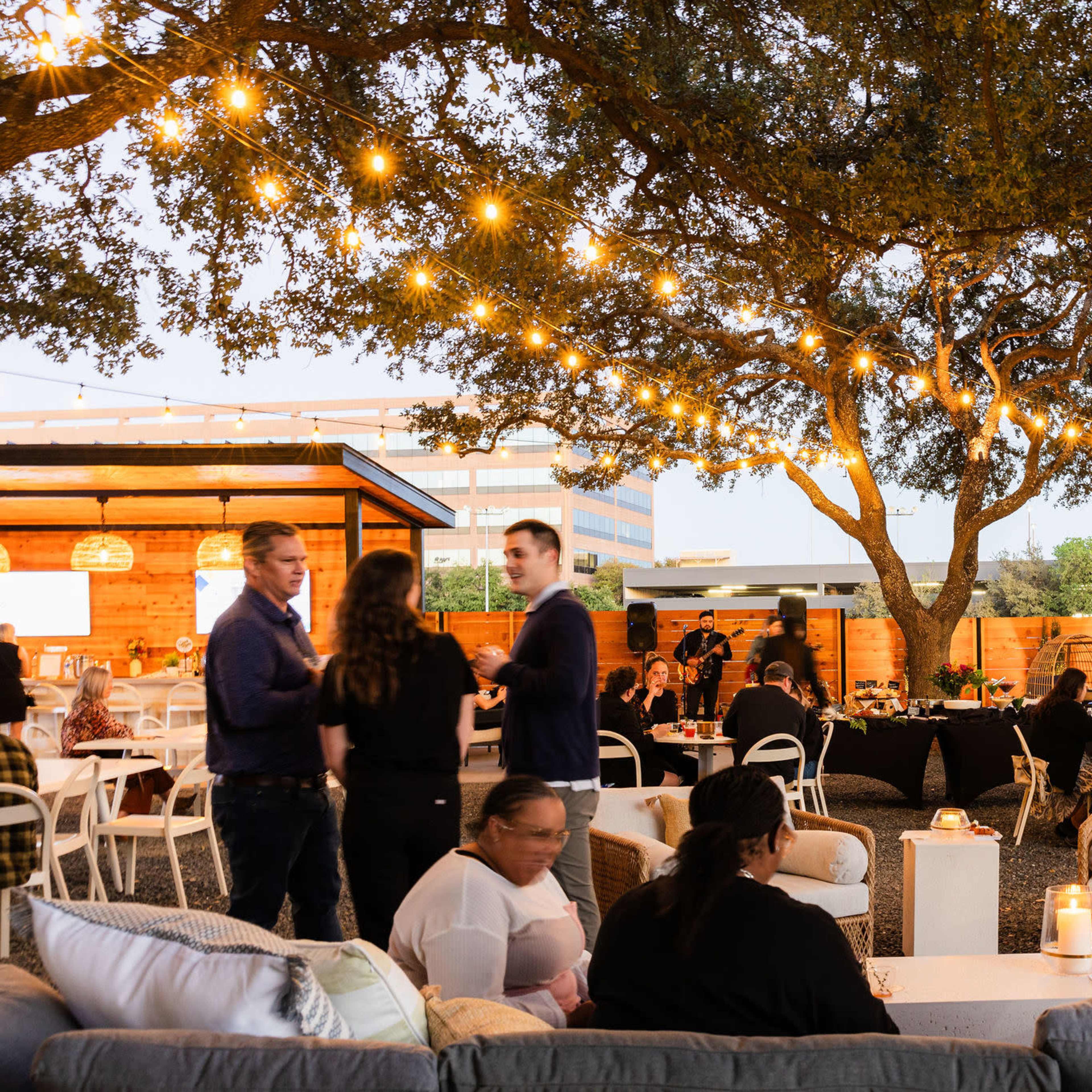 People socialize under string lights hanging from trees in an outdoor event space with seating and tables.