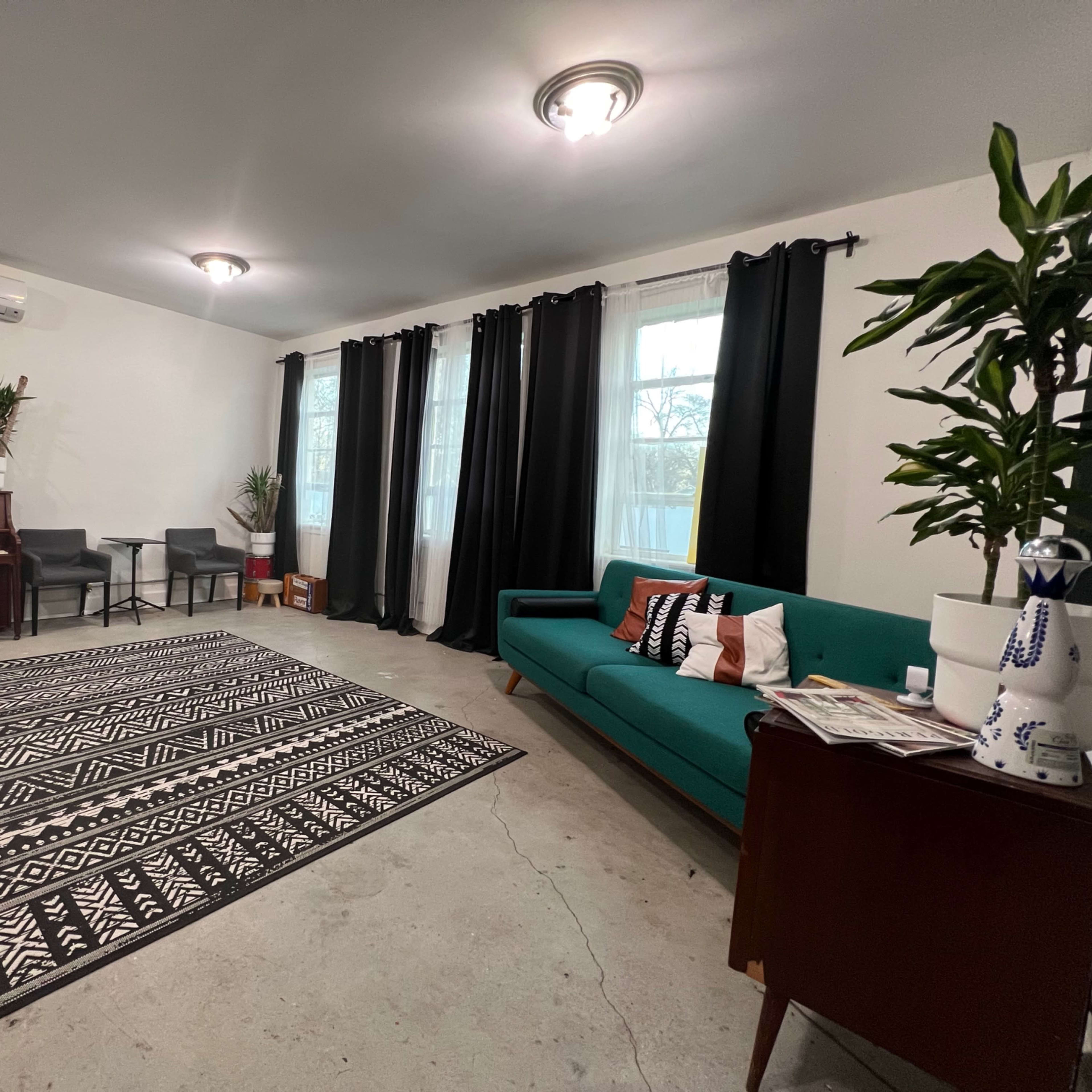 A spacious indoor living area featuring a piano, a green couch, a black and white patterned rug, and large windows with dark curtains.