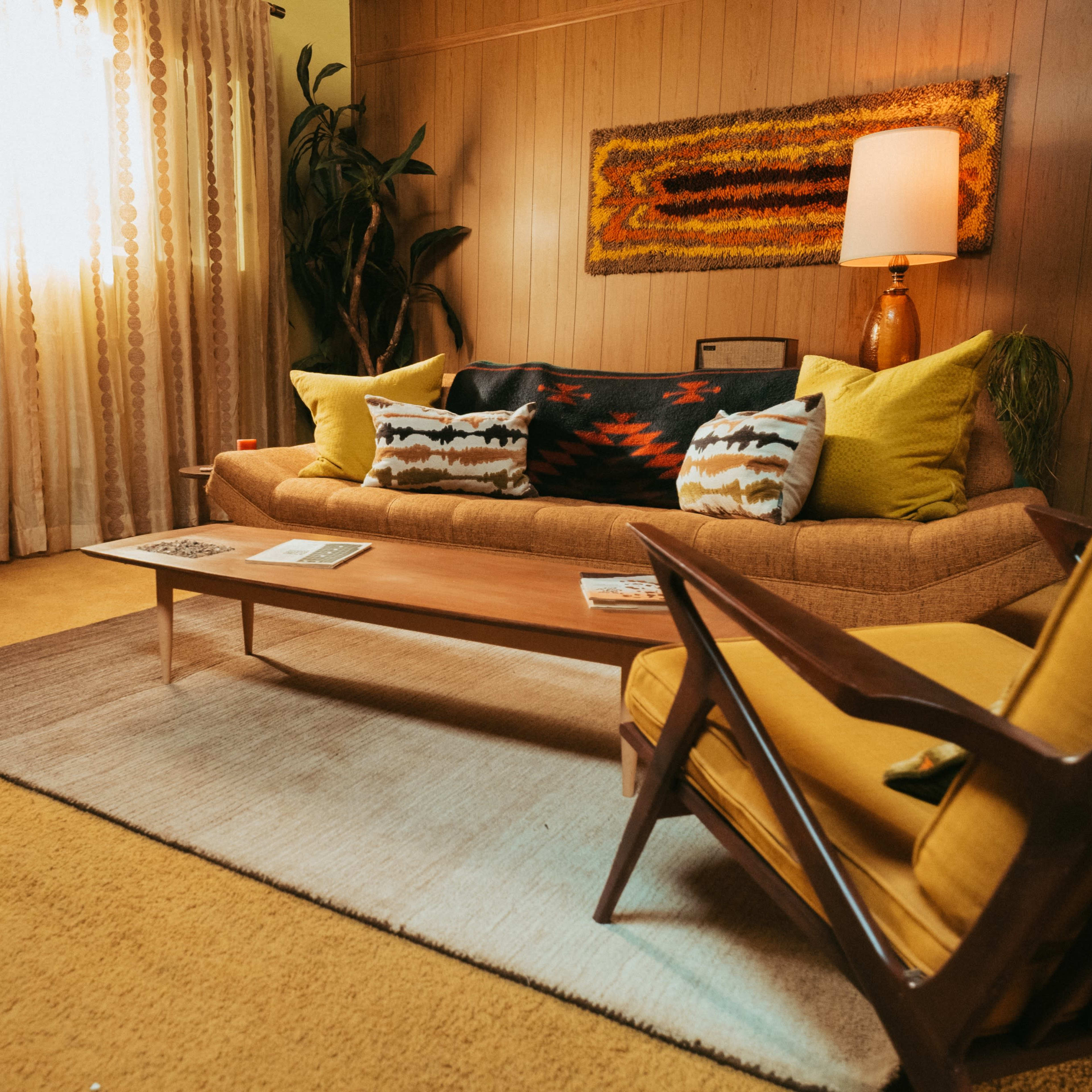 A vintage living room featuring a sofa with colorful cushions, a wooden coffee table, and a retro television set beside a large window with light curtains.