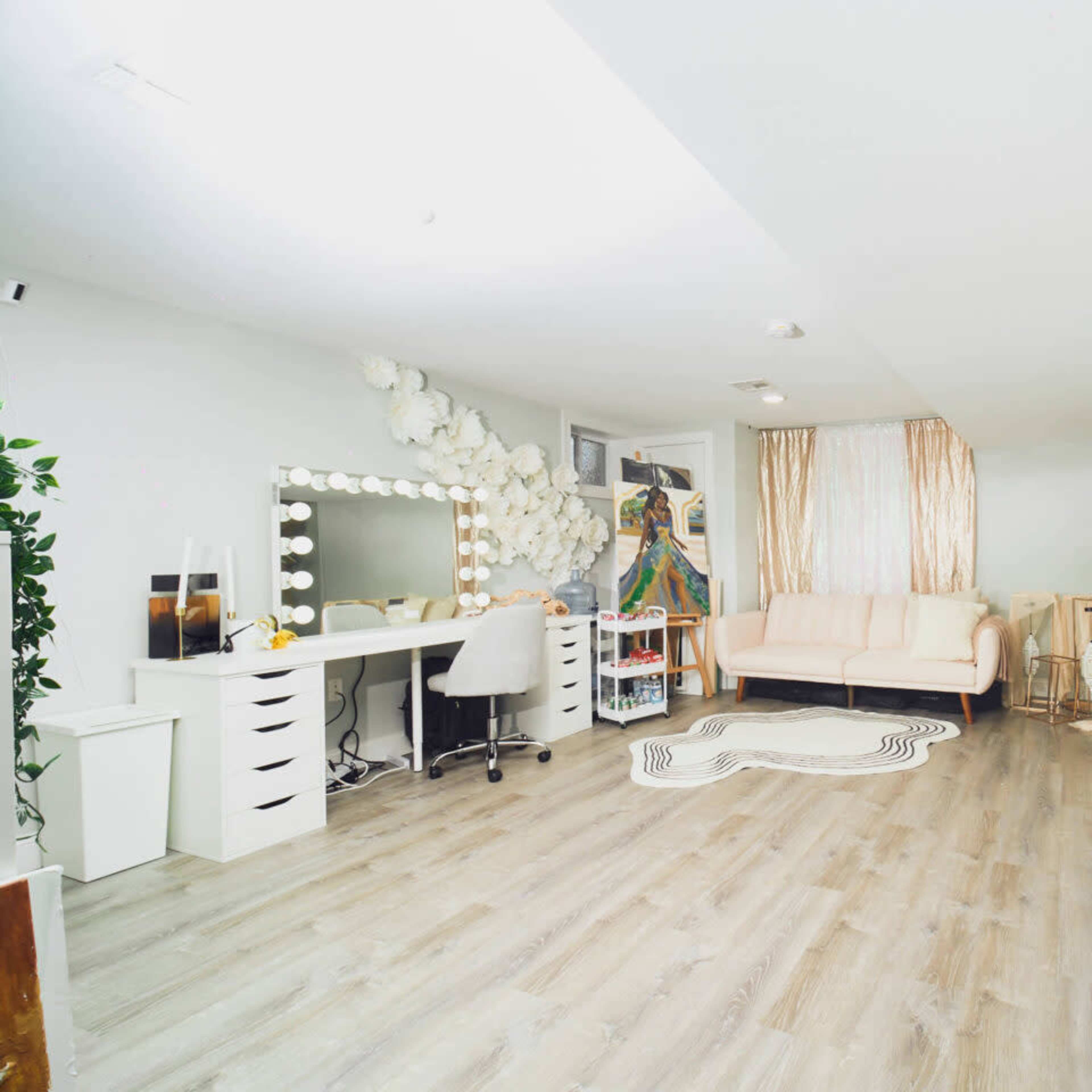 A modern, well-lit beauty studio with two large vanity stations, a cozy sofa, and decorative greenery on one wall.
