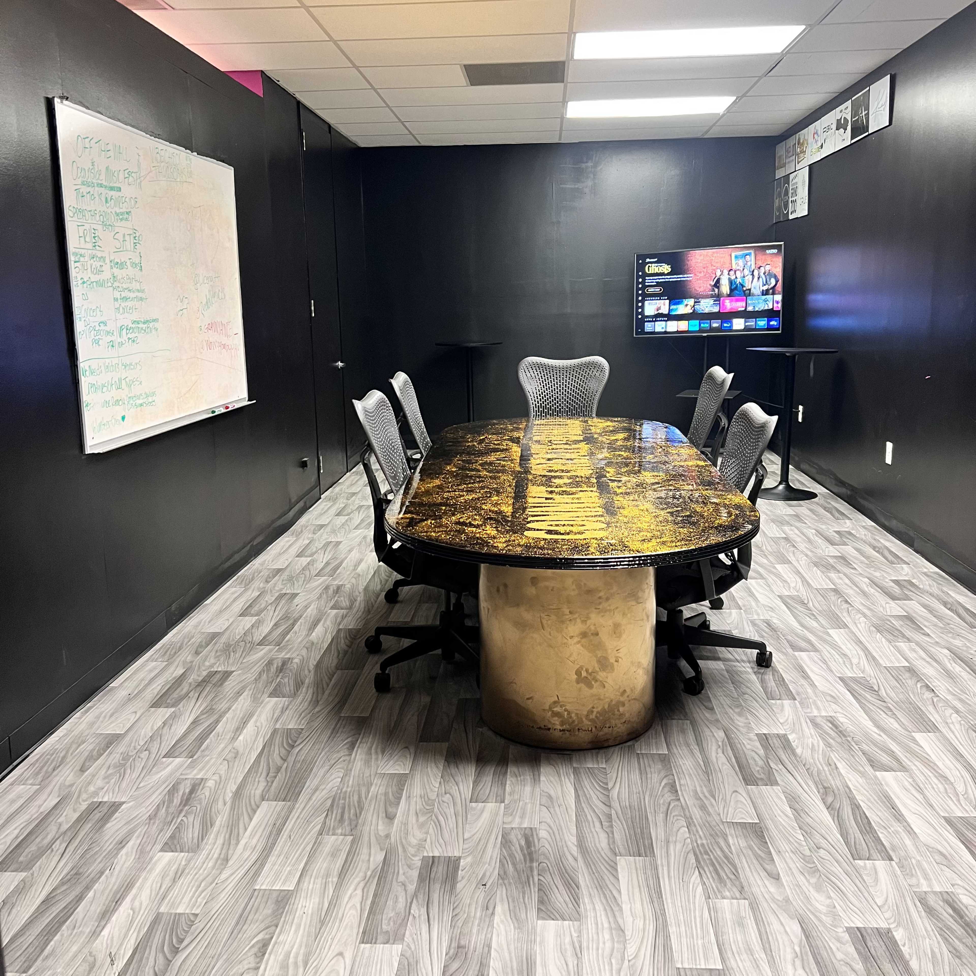 A conference room with a large oval table surrounded by chairs, a whiteboard on one wall, and a mounted screen displaying digital content.