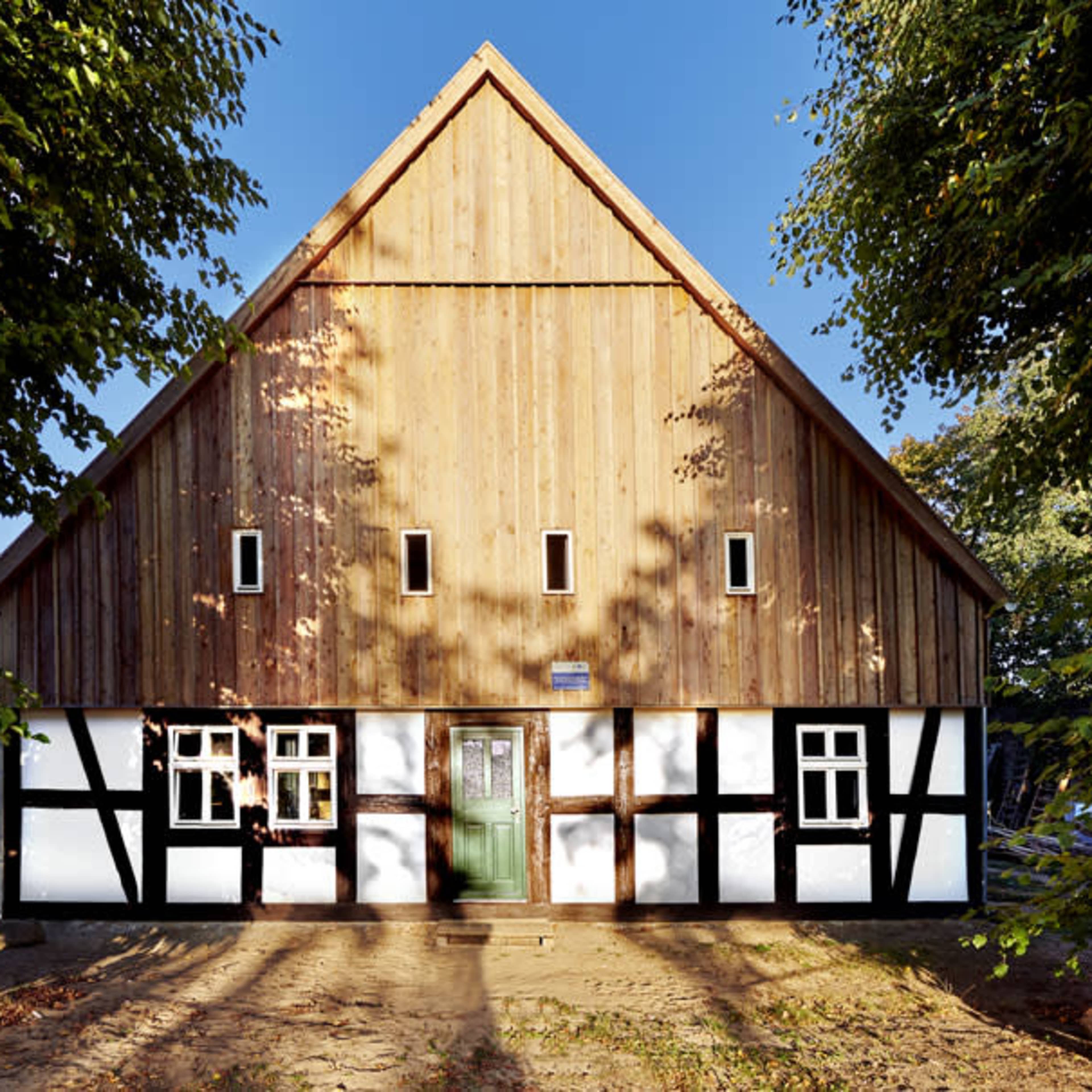 A traditional-style wooden house with a steeply pitched roof and a mix of wood and white plaster on the lower walls, surrounded by trees.