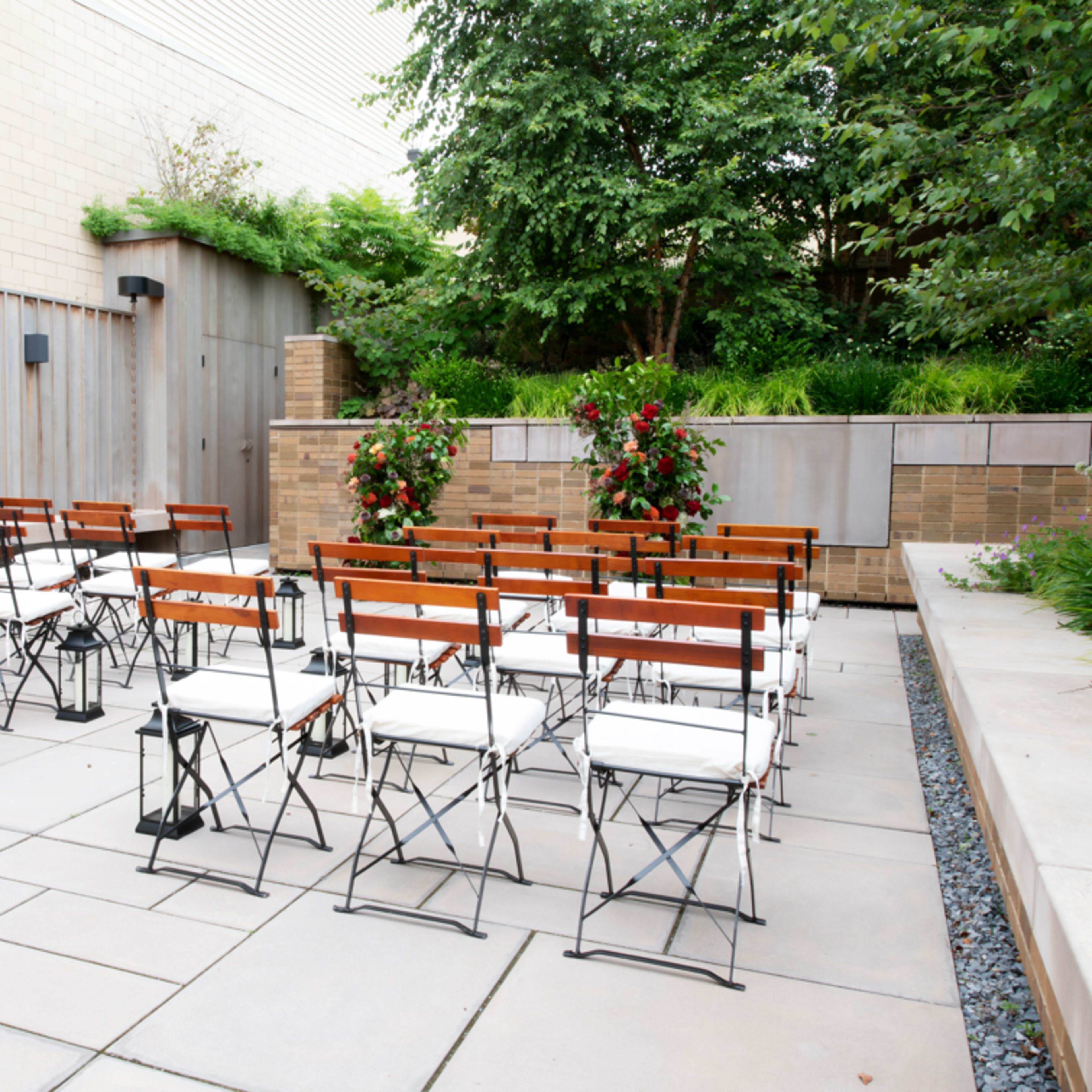 A set of wooden foldable chairs arranged in rows on a paved outdoor area surrounded by greenery.