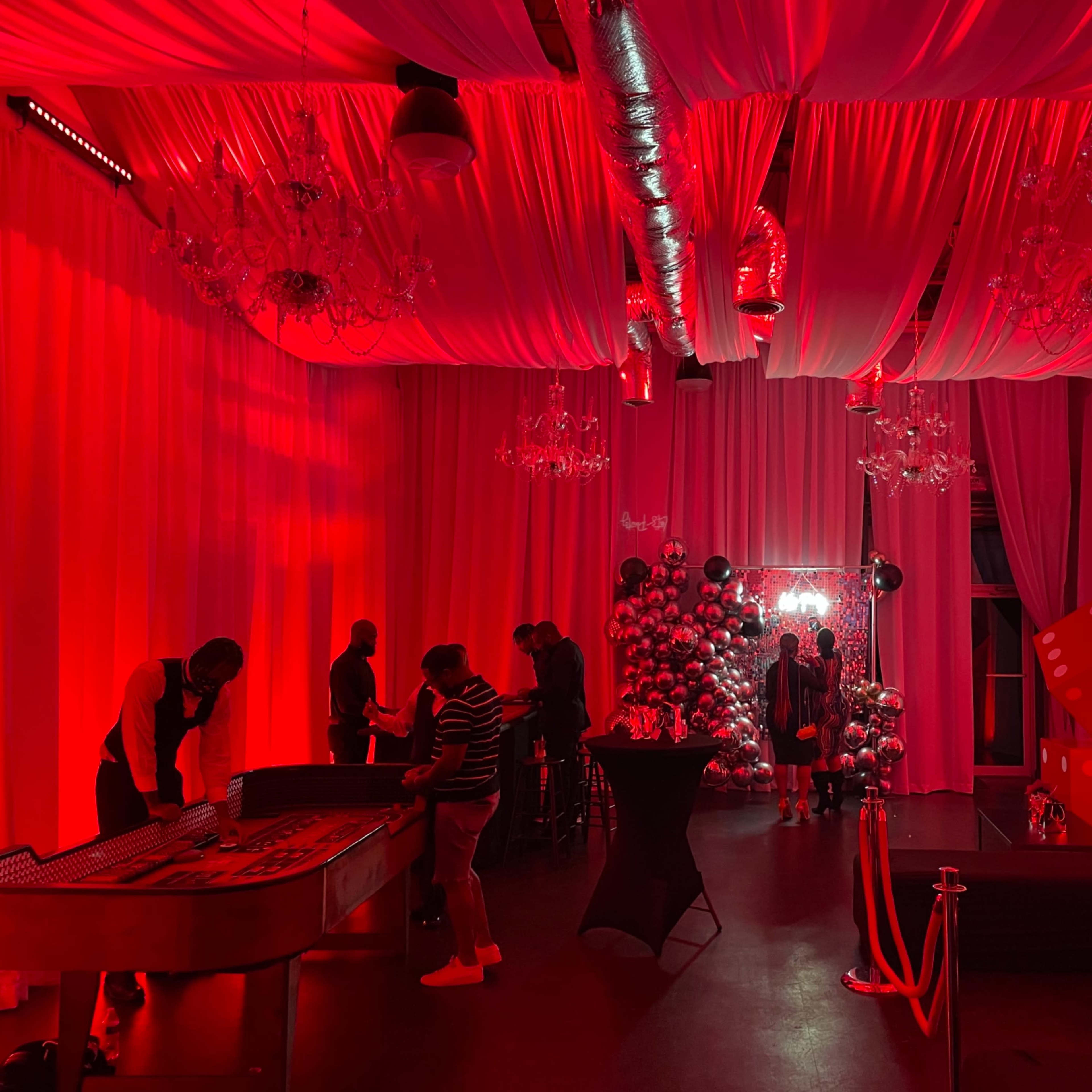 A dimly lit event space decorated with red lighting, elegant chandeliers, and a Christmas tree surrounded by festive decorations, with guests mingling and playing games.