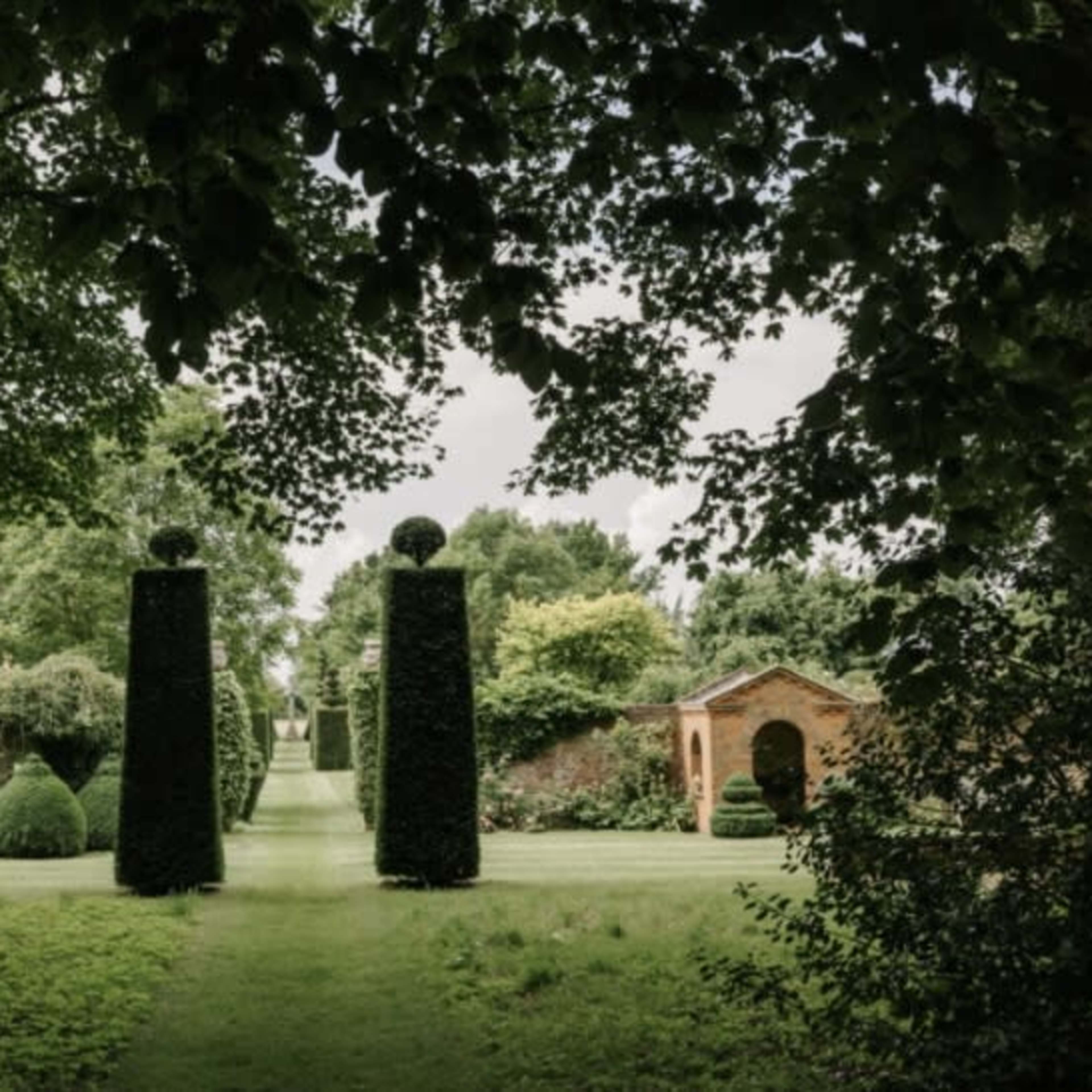 A landscaped garden featuring tall, neatly trimmed hedges and symmetrical trees flanking a pathway that leads to a brick structure in the background.