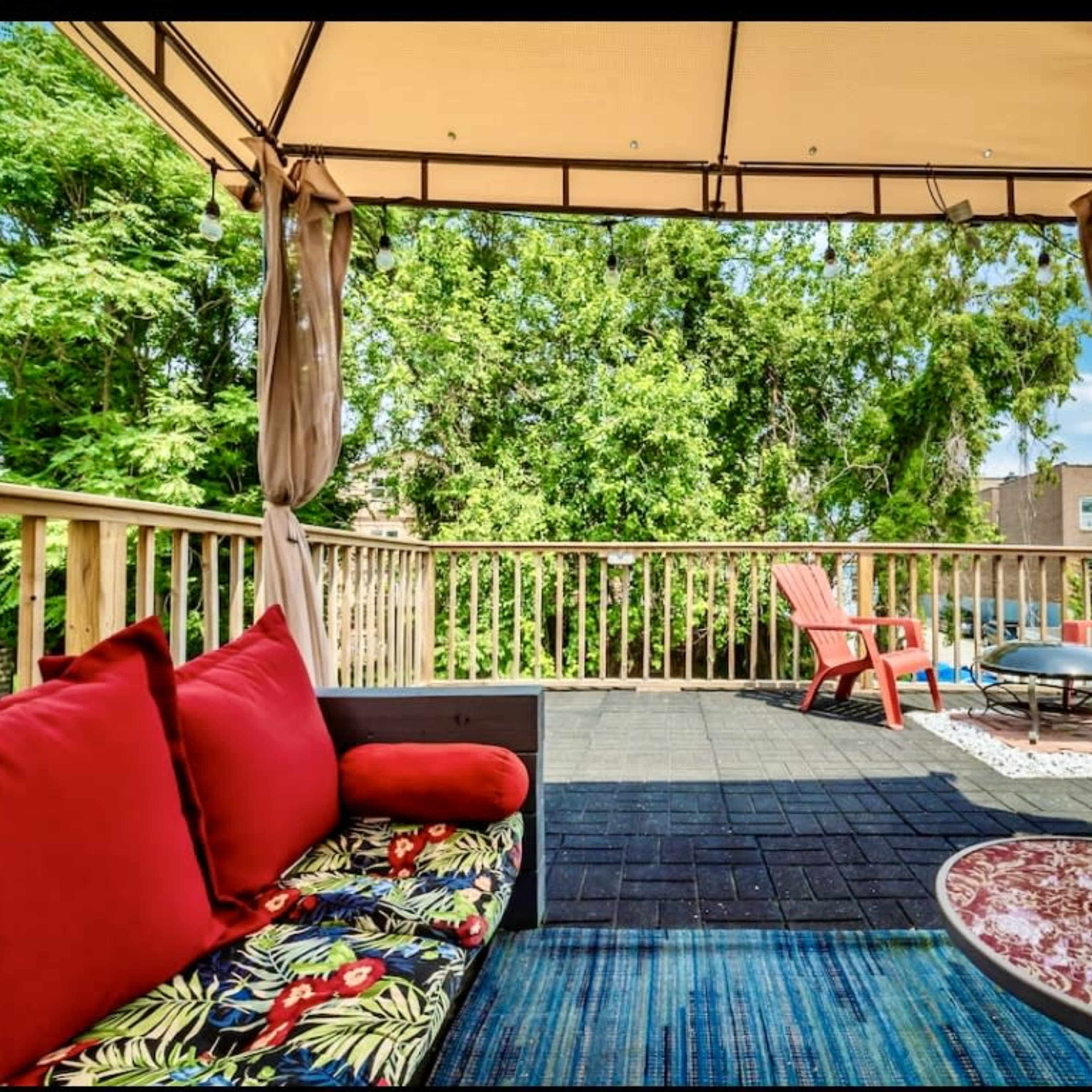 A patio featuring a gazebo with draped curtains, colorful seating options, and a view of trees.