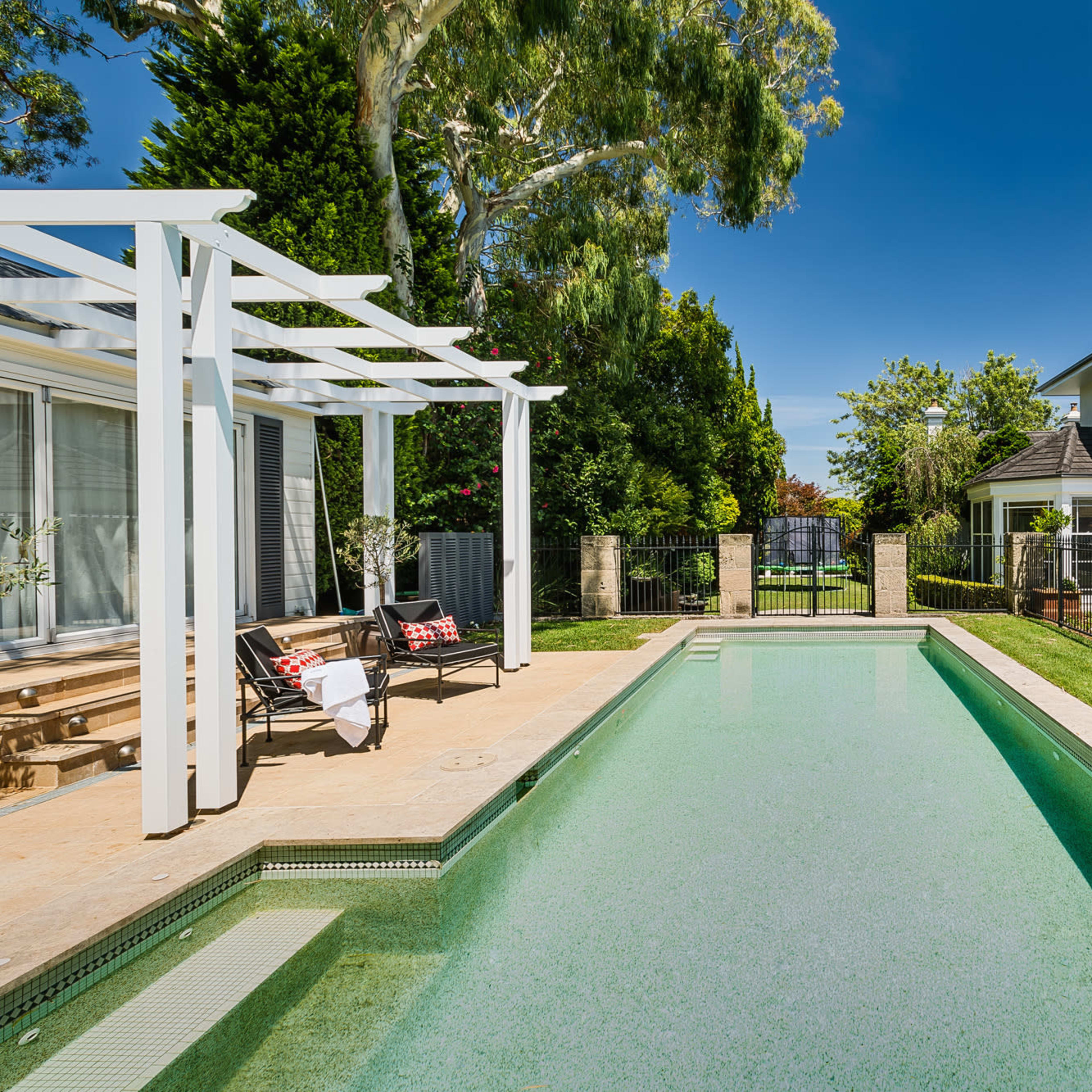 A clear swimming pool surrounded by a well-maintained garden and a two-story house in the background.