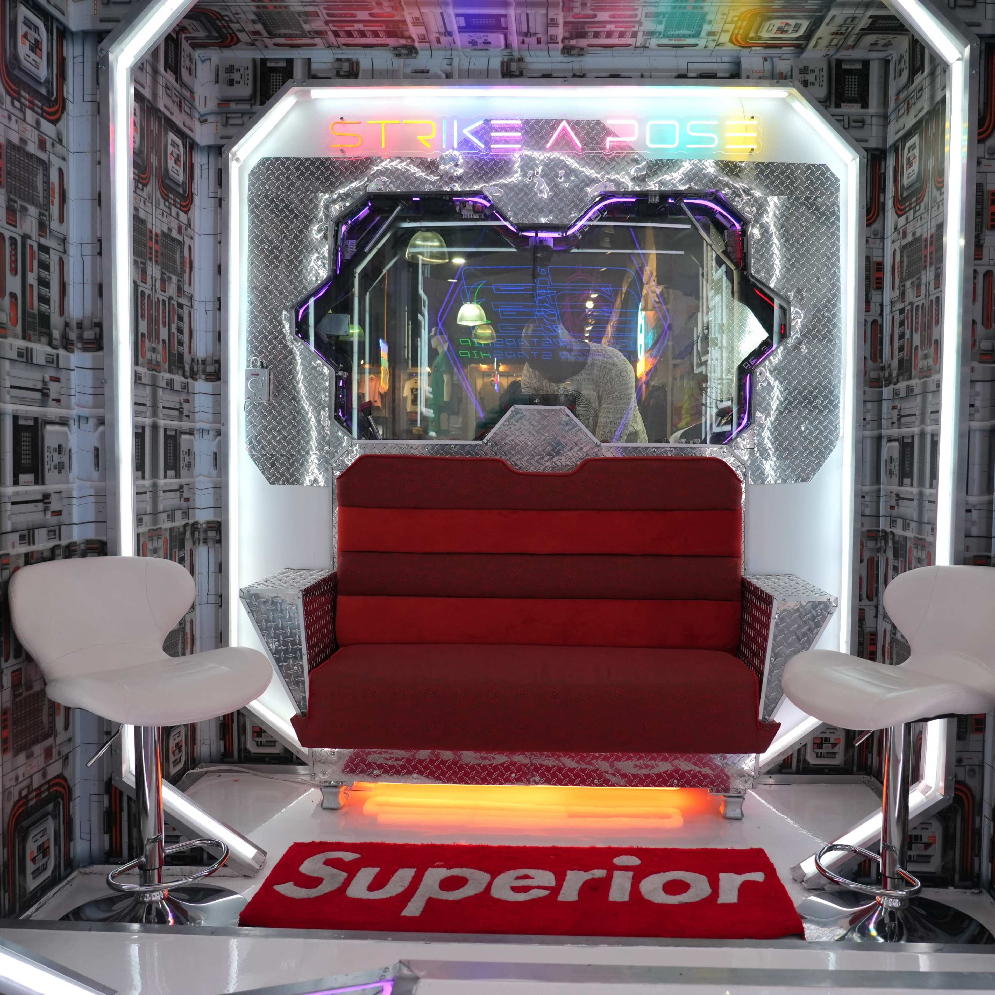 A futuristic photo booth setup with a red sofa and two white chairs surrounded by patterned walls and colorful lighting.