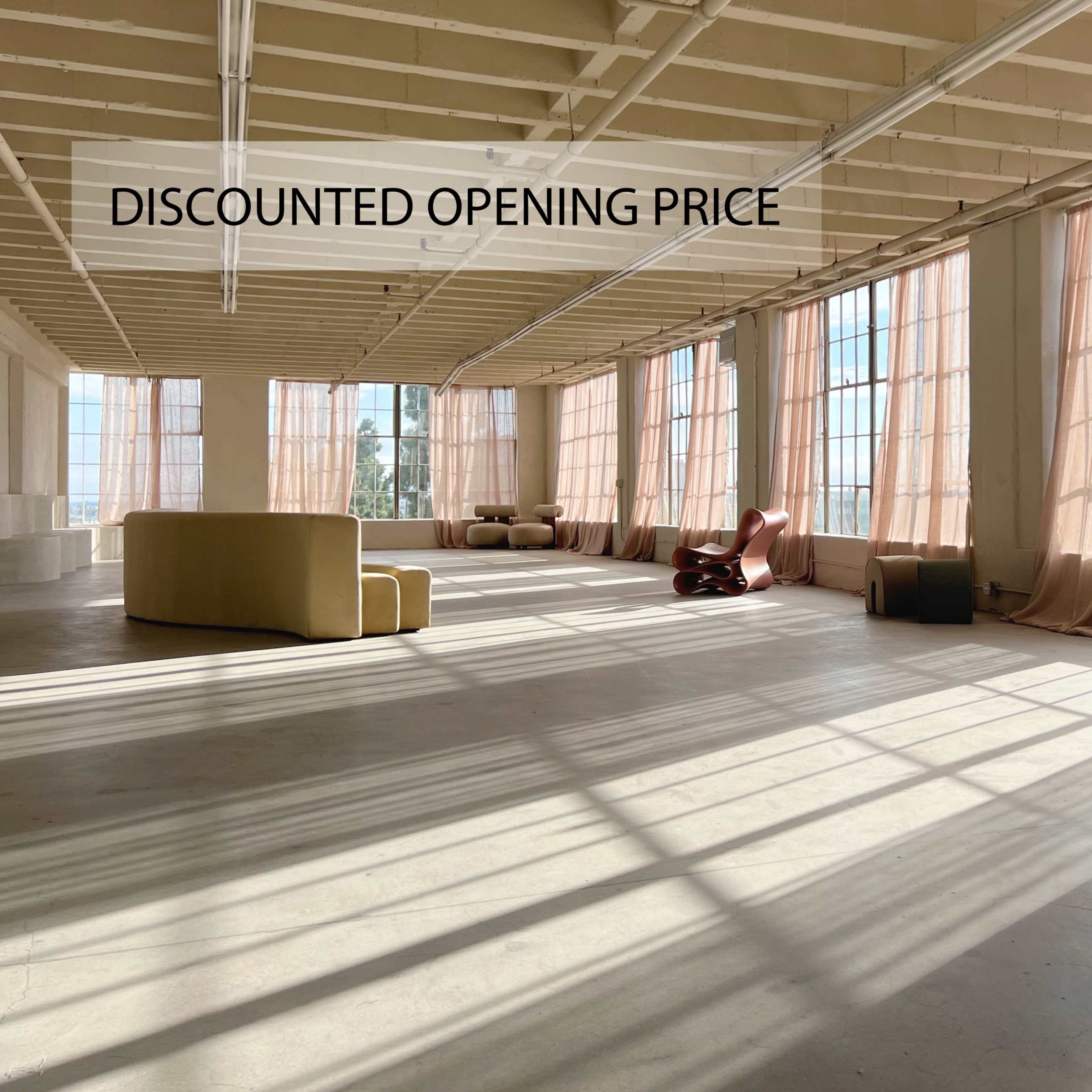 A spacious, well-lit room features large windows with sheer curtains, showcasing minimalist furniture and light streaming across the floor.