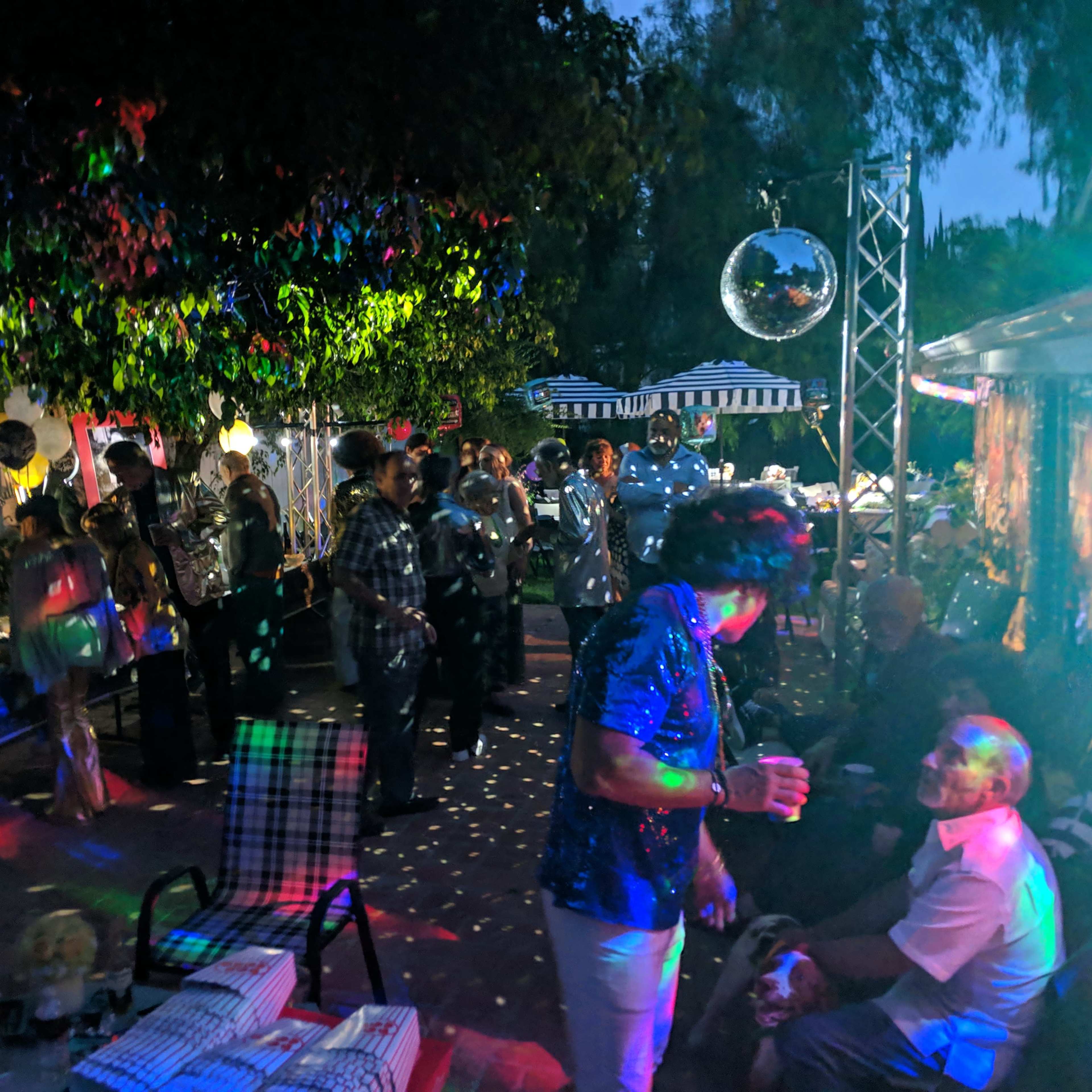 A group of people dances under colorful lights at an evening gathering in a garden setting.