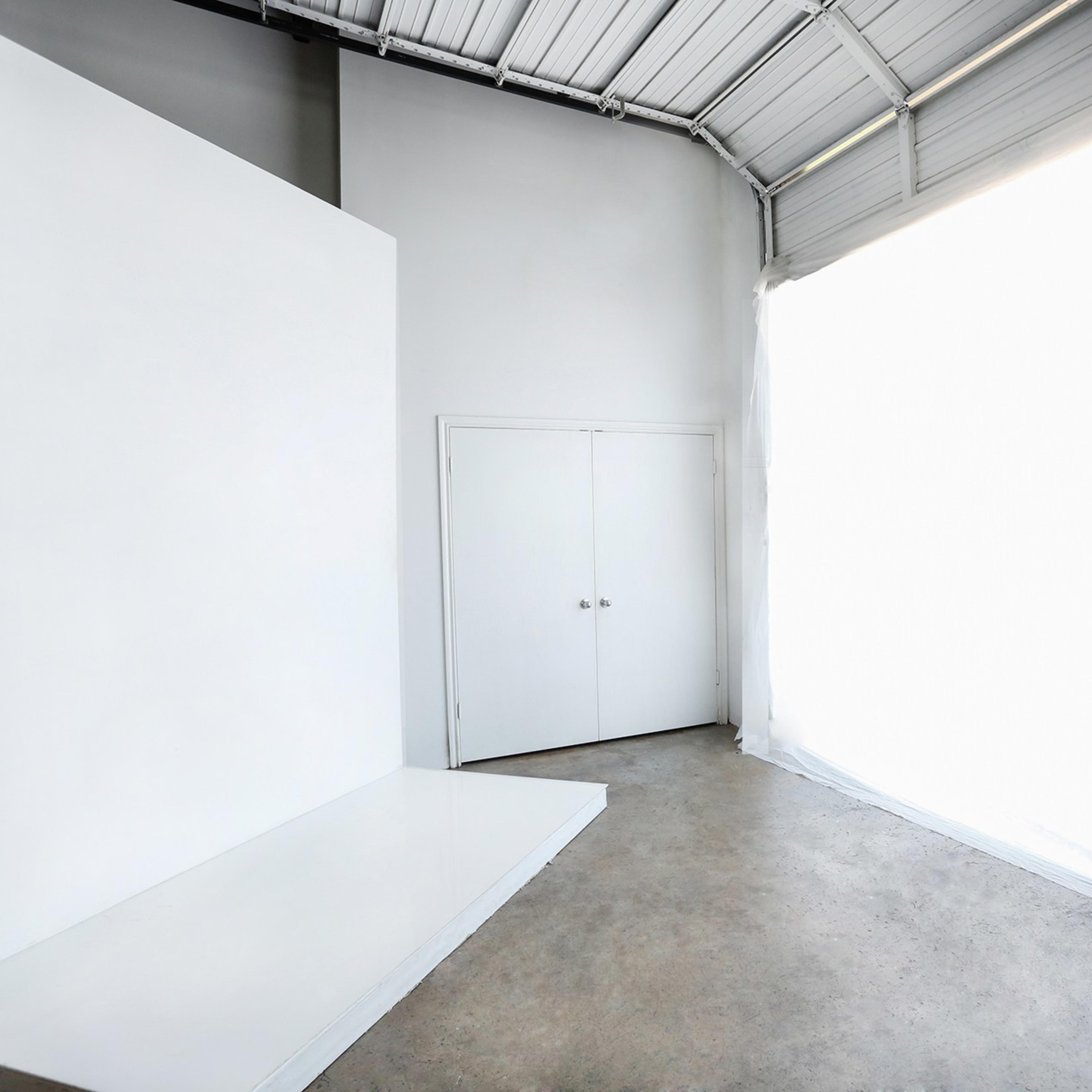A spacious, minimalist room features a large white backdrop and a double door on one wall, with natural light flowing in from an expansive window.