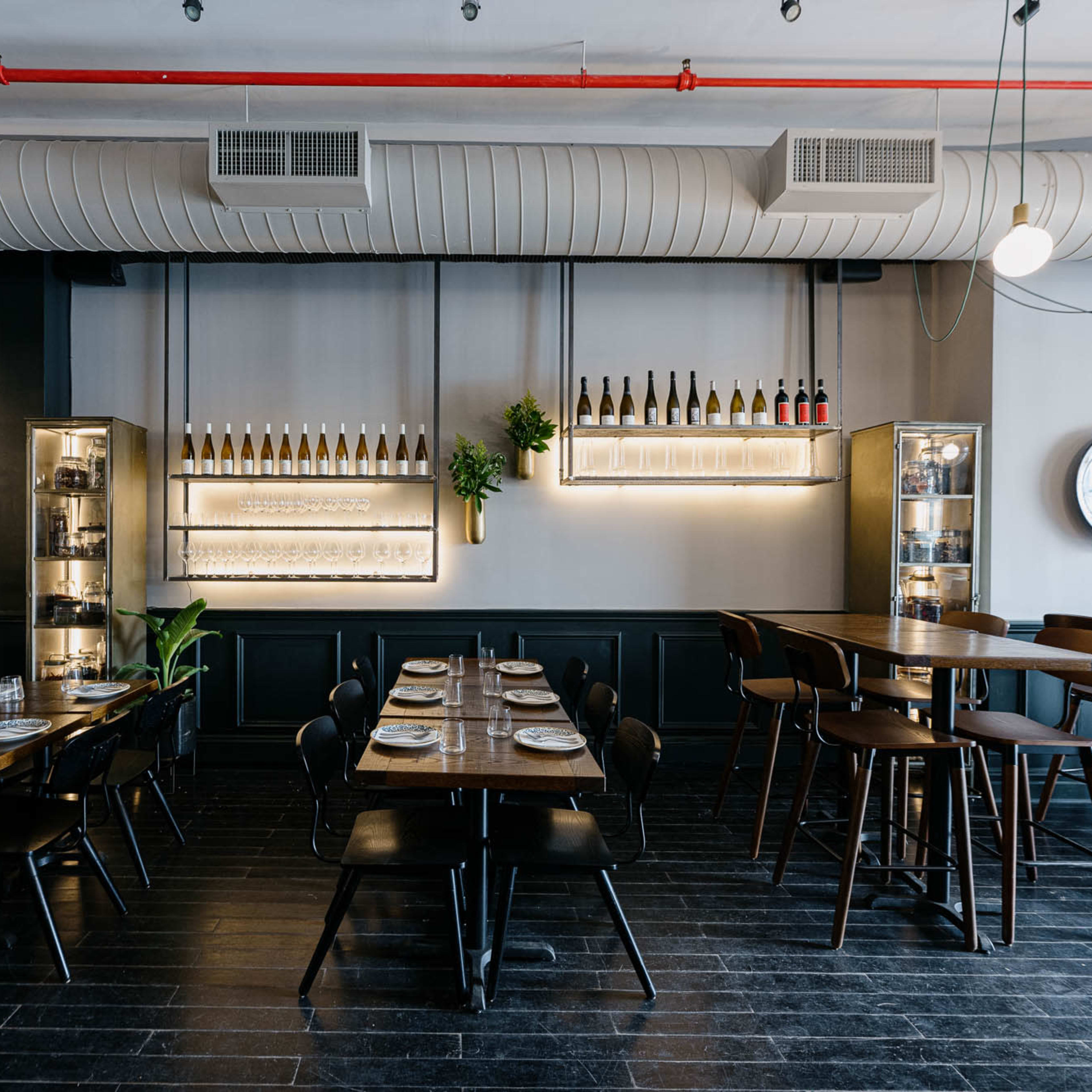A modern restaurant interior with wooden tables, black chairs, and a wall displaying neatly arranged bottles.