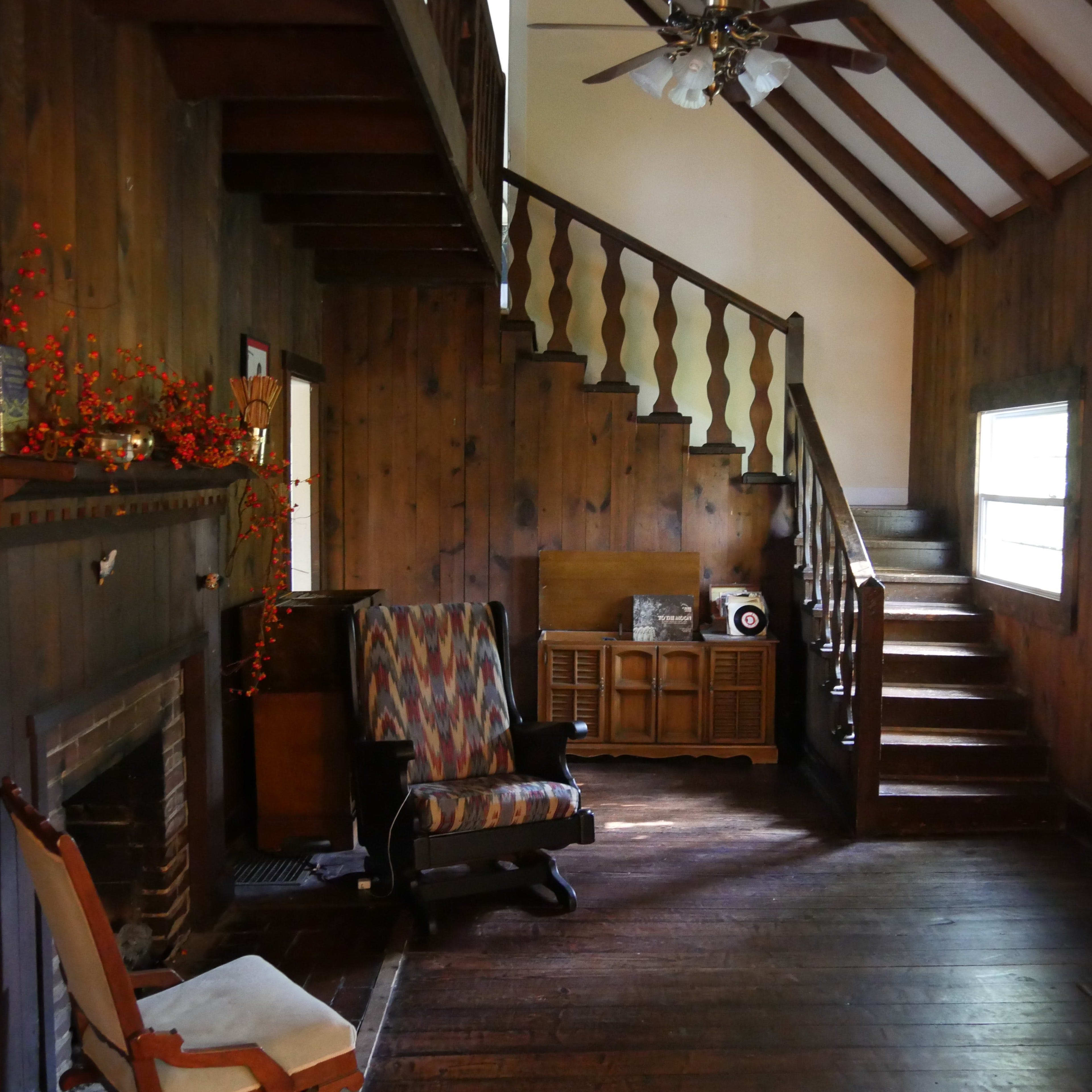 The interior of a wooden-paneled living room with a staircase, a fireplace, and a patterned armchair.