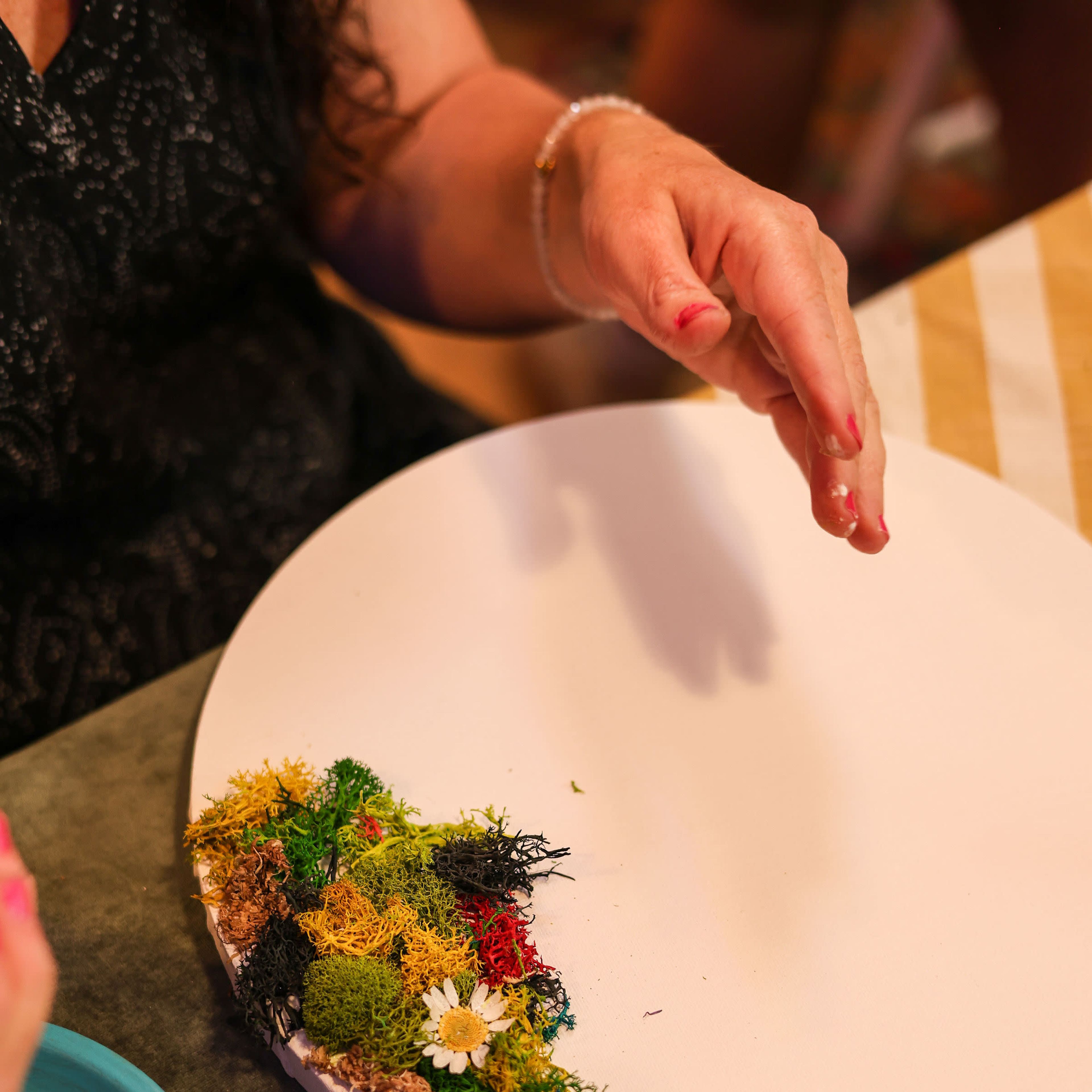 A person arranges colorful moss and small flowers on a white circular plate.