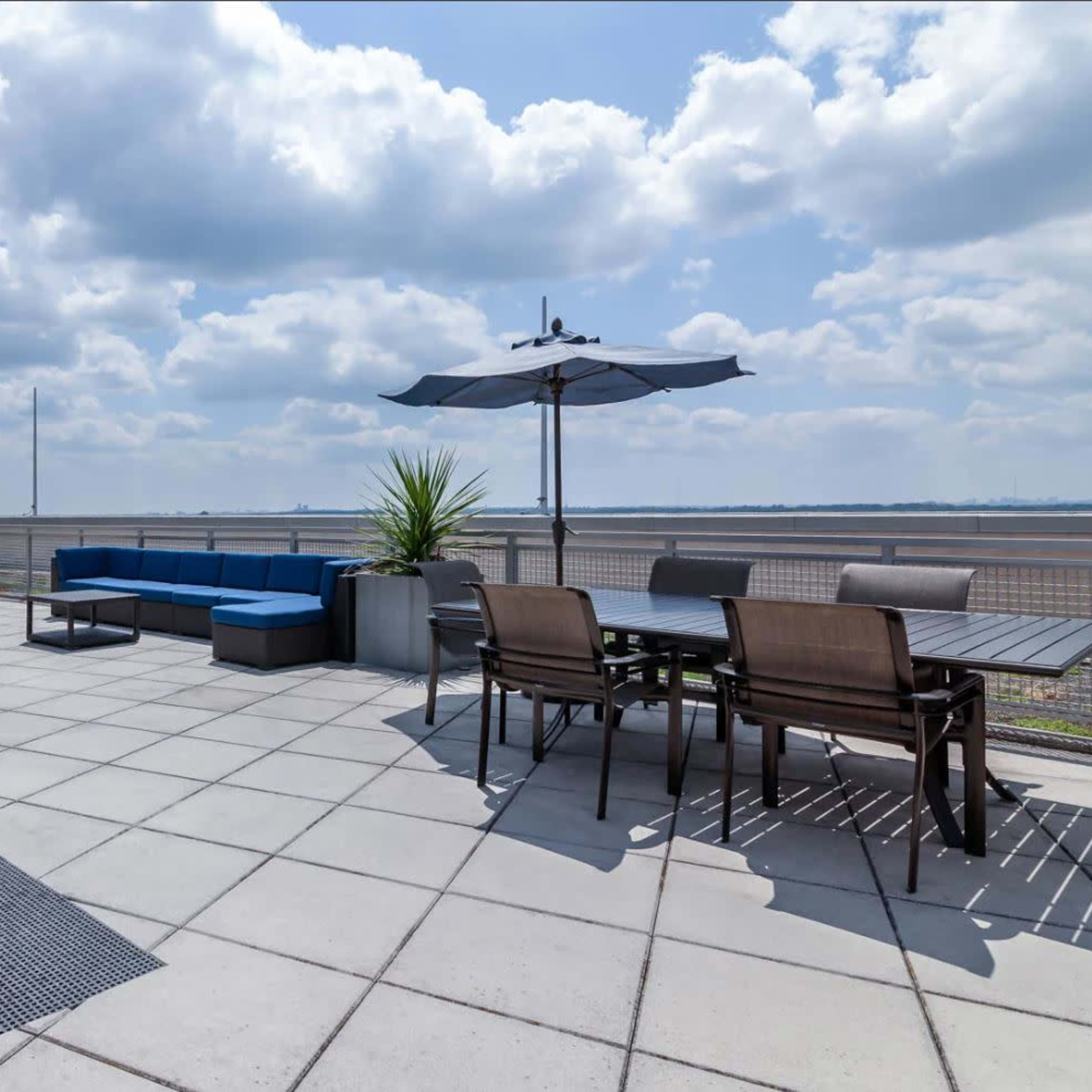 A spacious rooftop terrace features seating areas with furniture, a grill, and an umbrella overlooking a distant body of water.