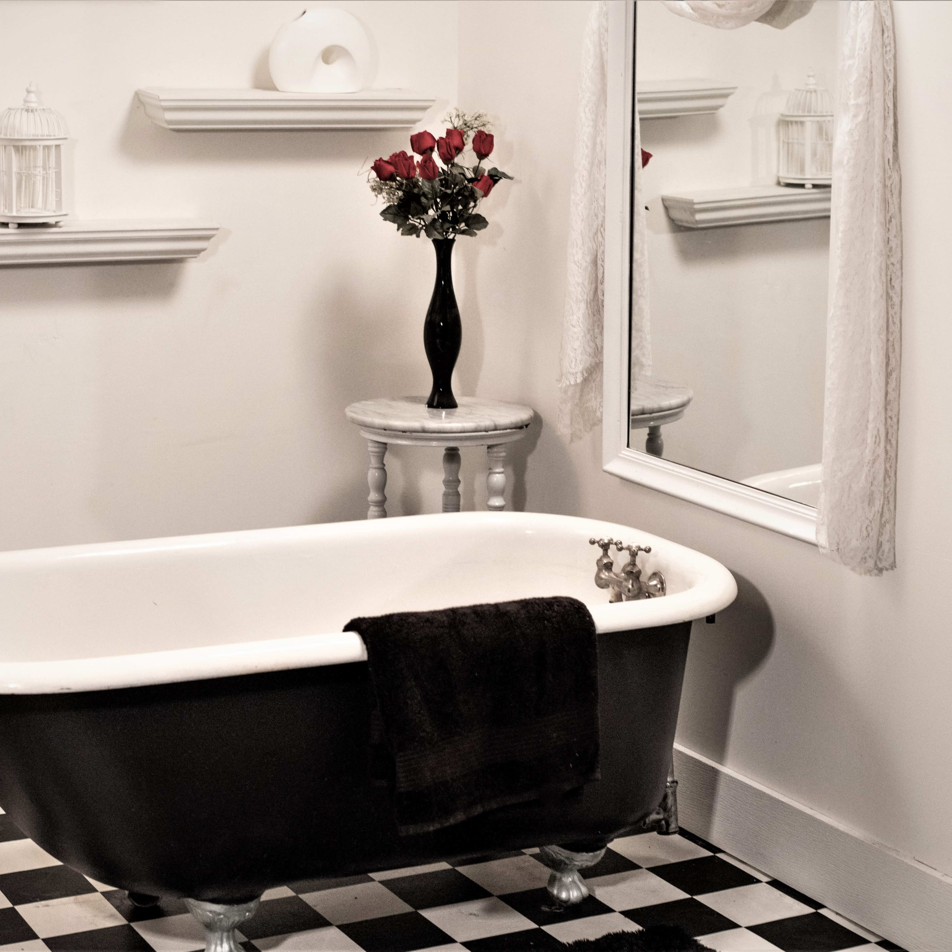 A black and white checkered bathroom features a vintage clawfoot bathtub, a vase of roses on a small table, and a wall mirror.