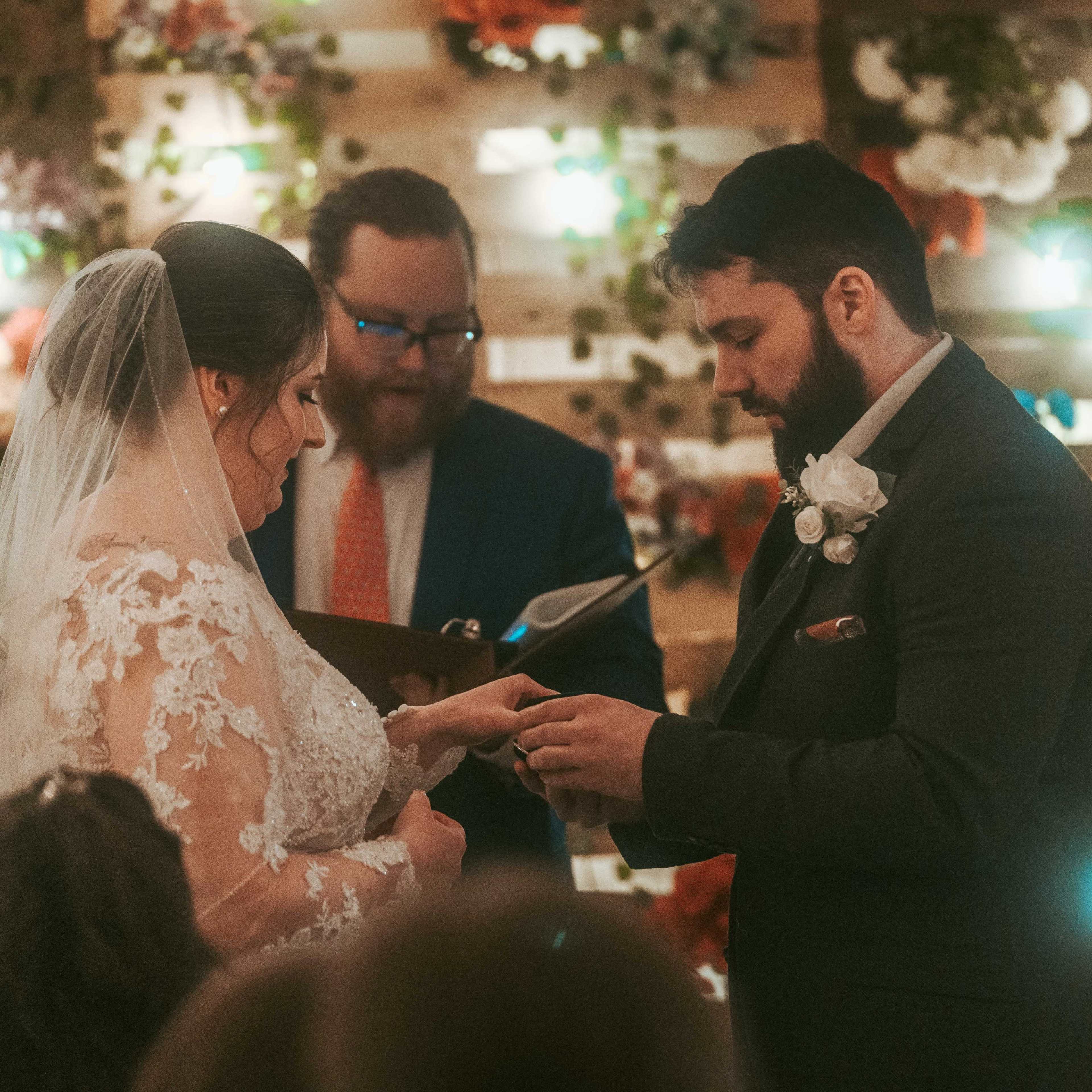 A couple exchanges wedding rings during their ceremony in front of an officiant amidst a decorated backdrop of flowers and lights.