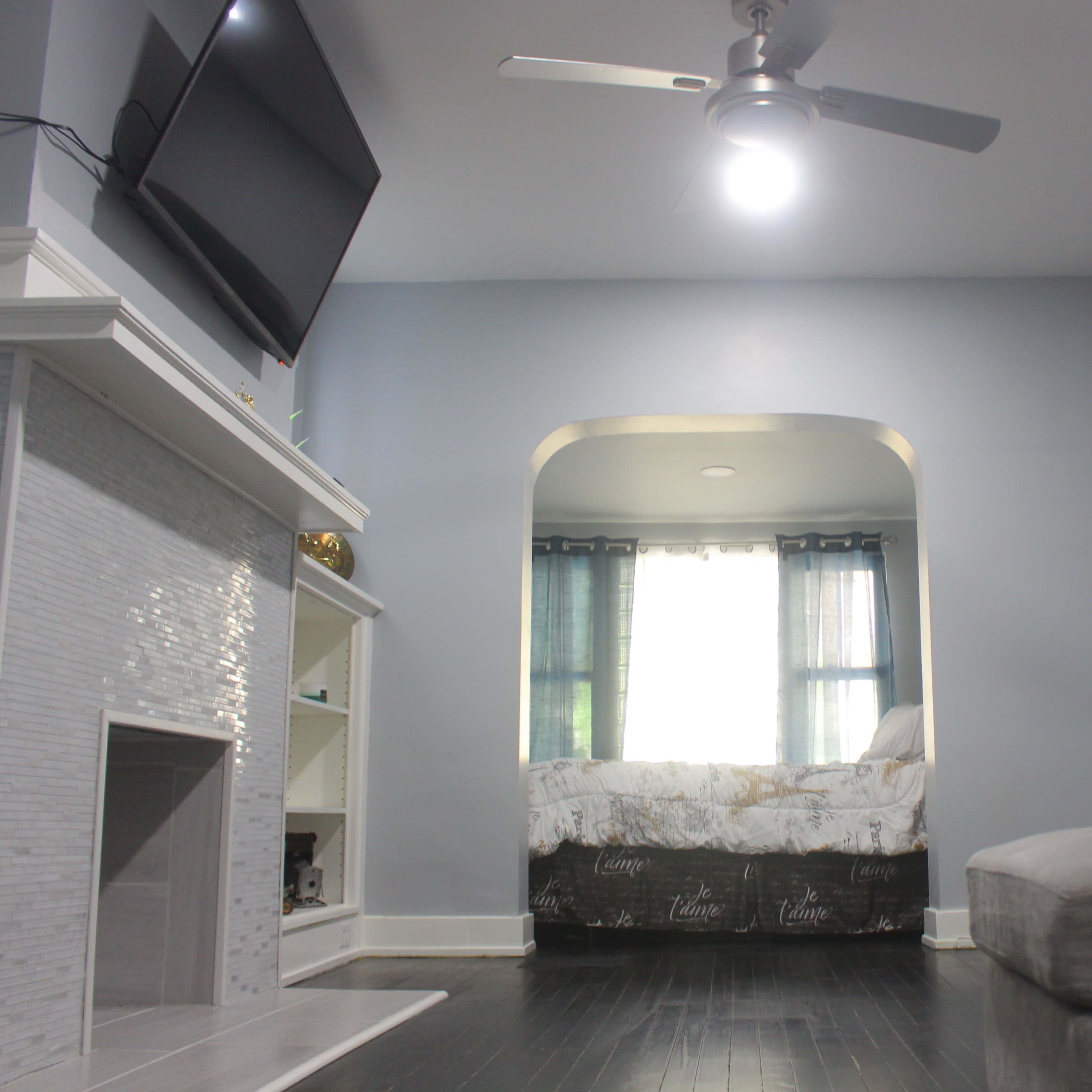 A living room with a light gray wall, a television mounted on the wall, a gray sectional sofa, and an archway leading to a bedroom with a bed and curtains.
