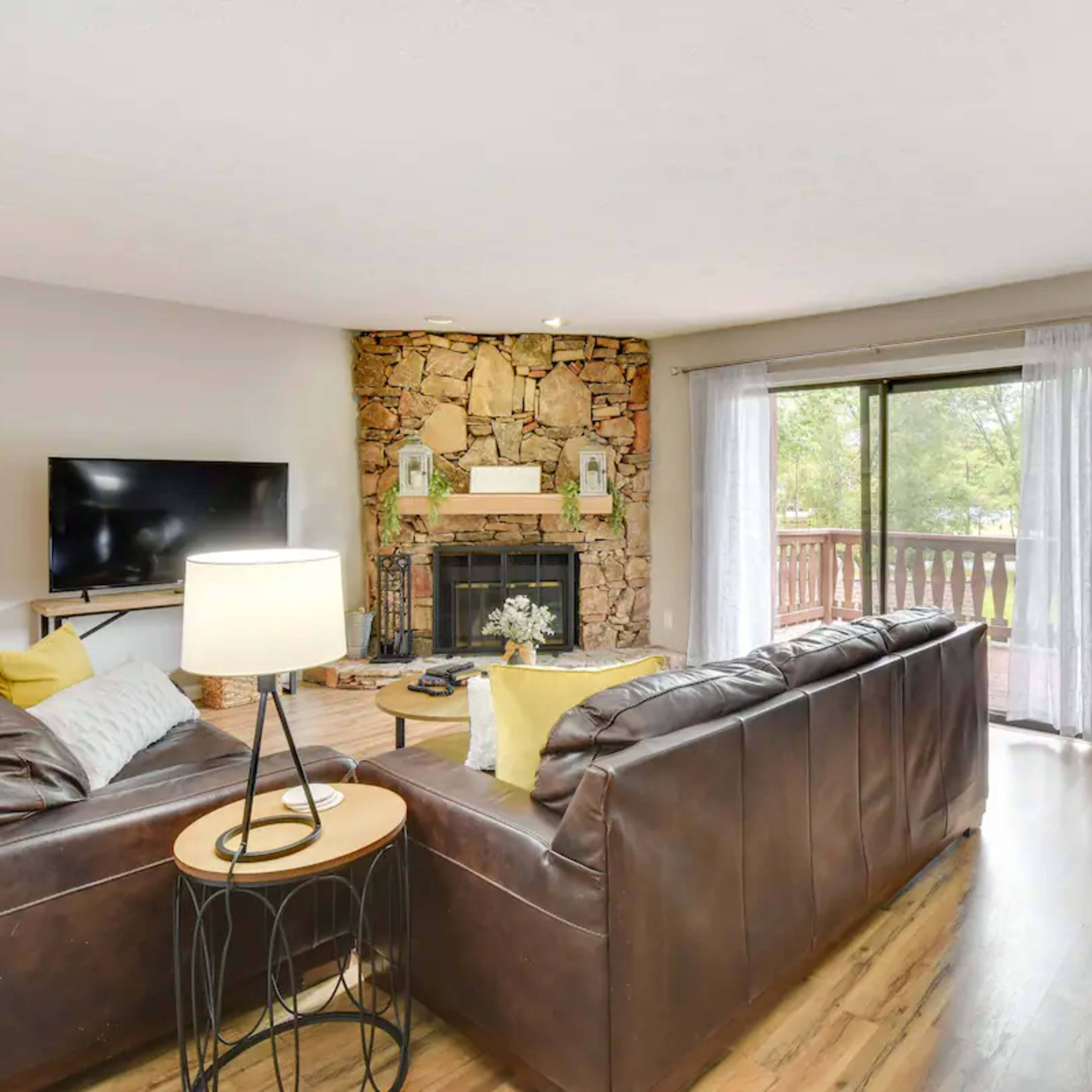 A living room with a stone fireplace, a dark leather sofa, a television, and large windows that open to a view of the outdoors.