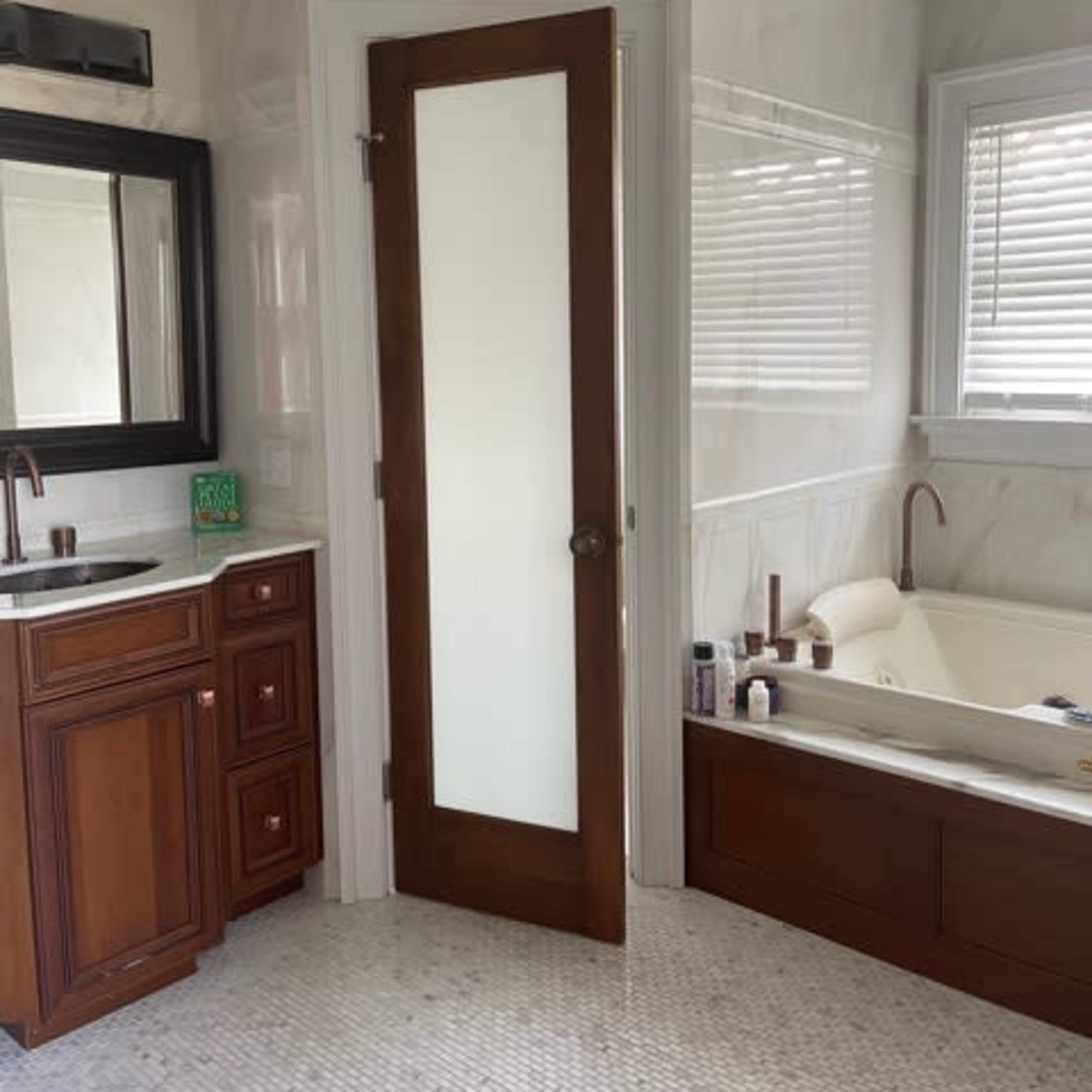 A modern bathroom features a wooden door with a glass panel, a marble-paneled wall, a vanity with brown cabinetry, and a bathtub beside a window.