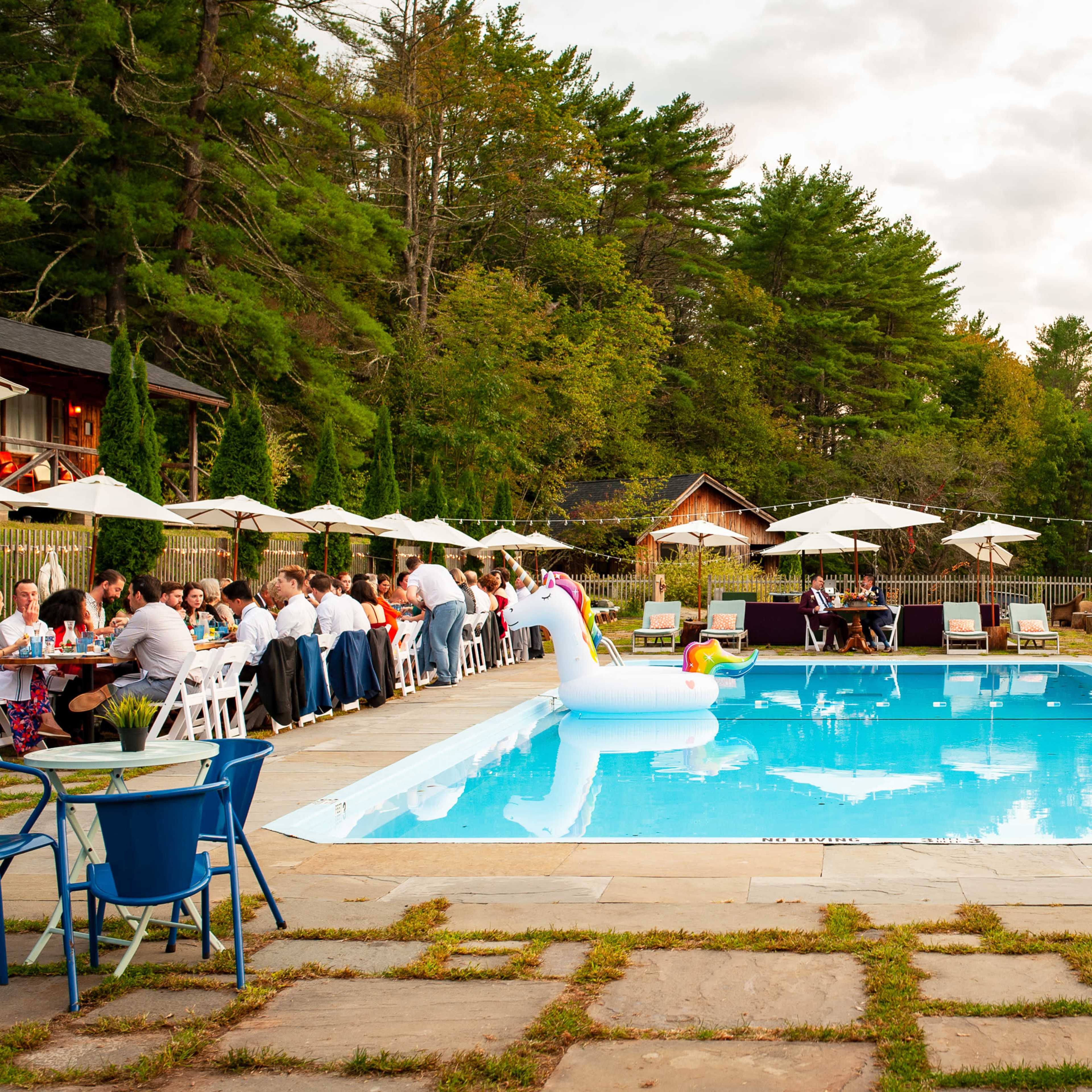 A large outdoor gathering is taking place around a swimming pool, with people seated at long tables and a colorful inflatable swan in the water.