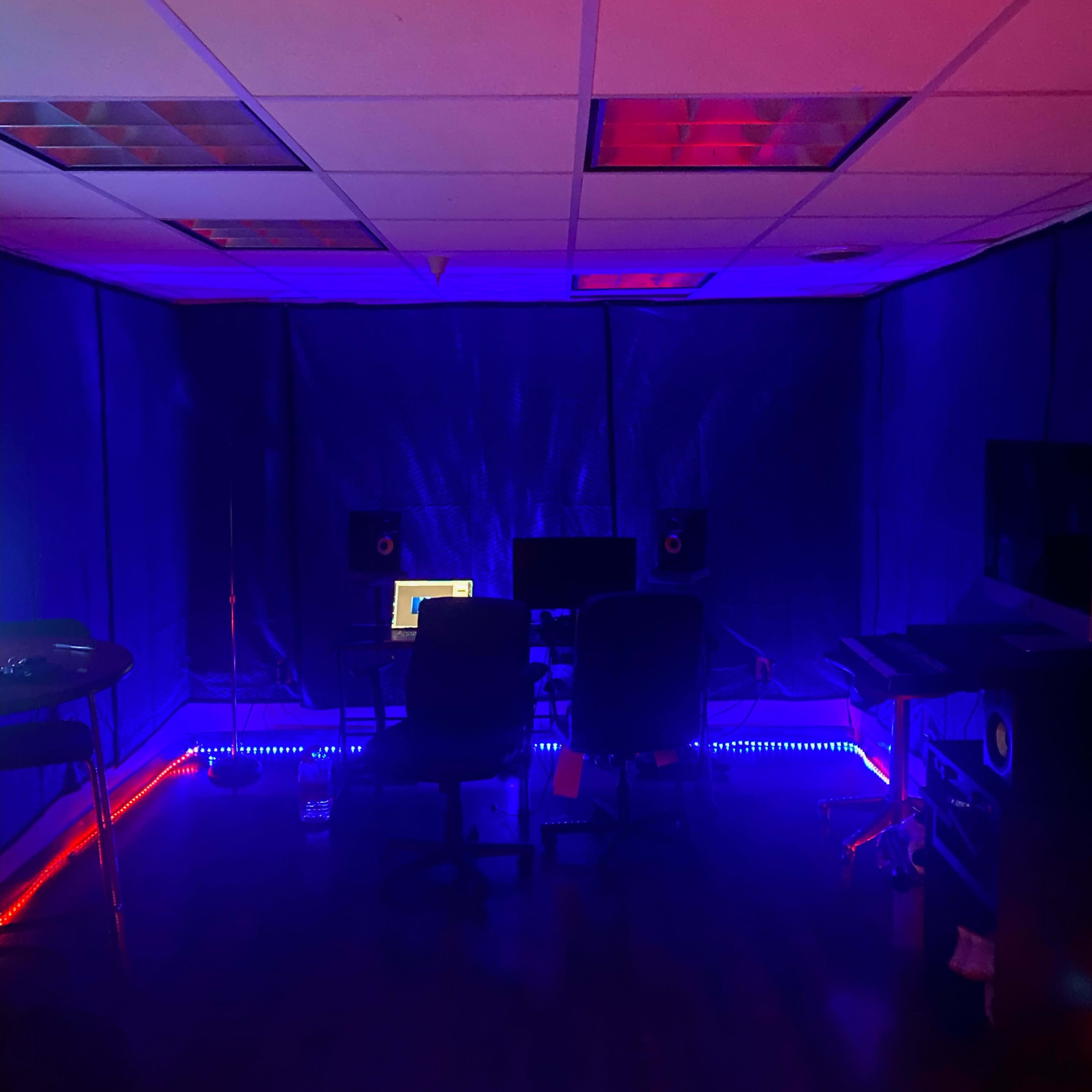 A dimly lit studio room with blue and red lighting, featuring two chairs and multiple computer screens set up on desks.