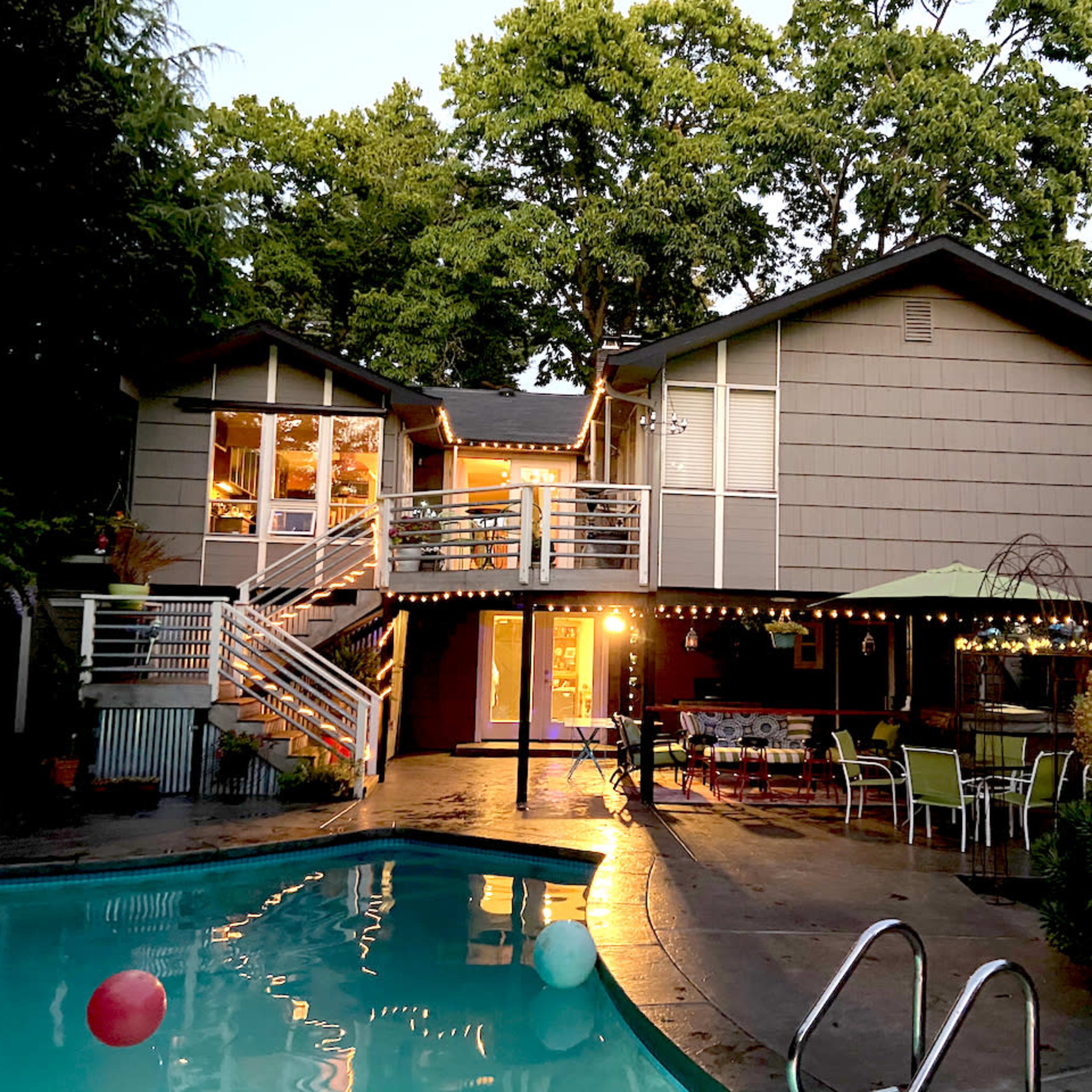 A two-story house with a patio and string lights, facing a swimming pool in the foreground.