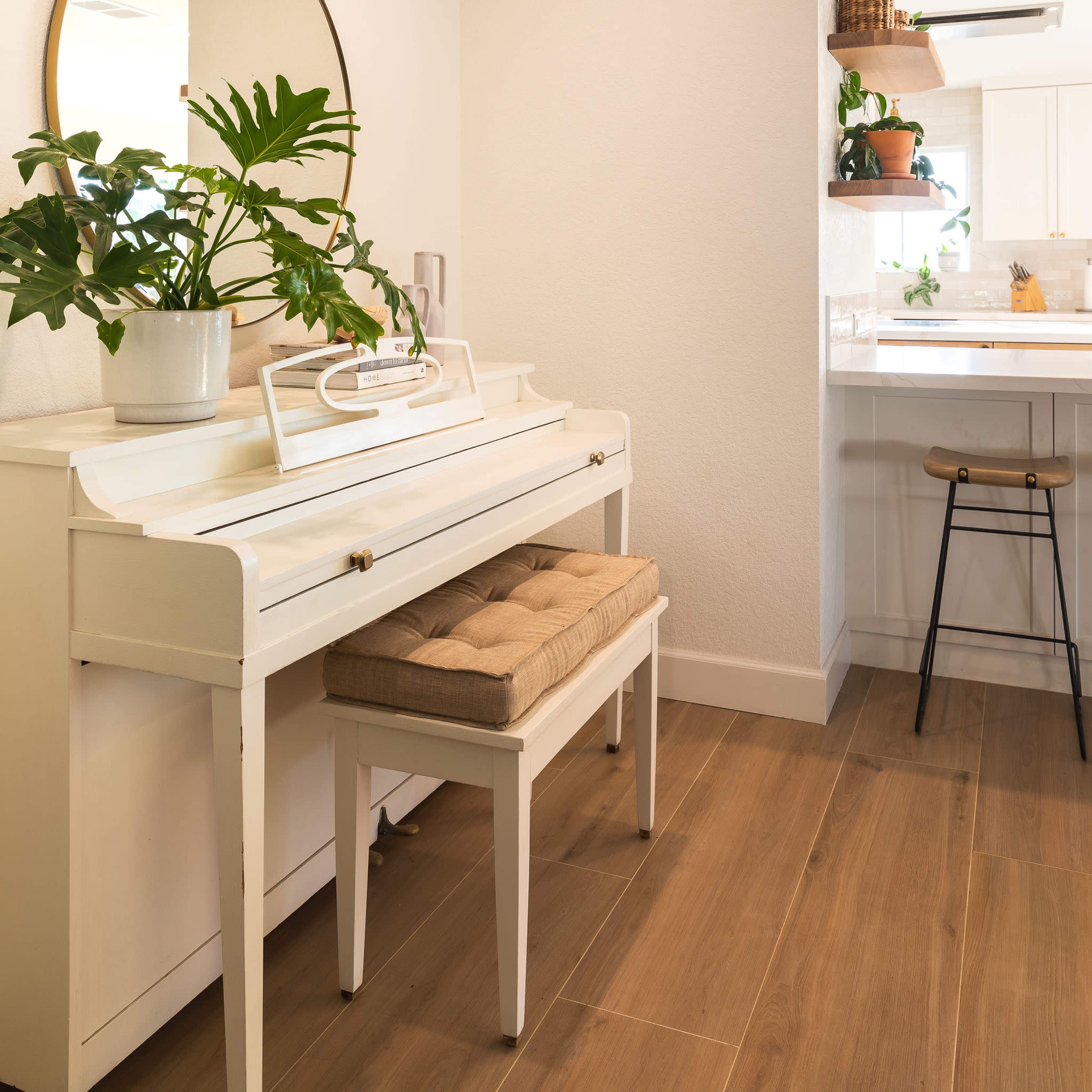 A white piano with a matching bench is positioned against a wall, accompanied by a round mirror and a potted plant, in a bright, well-lit entryway.