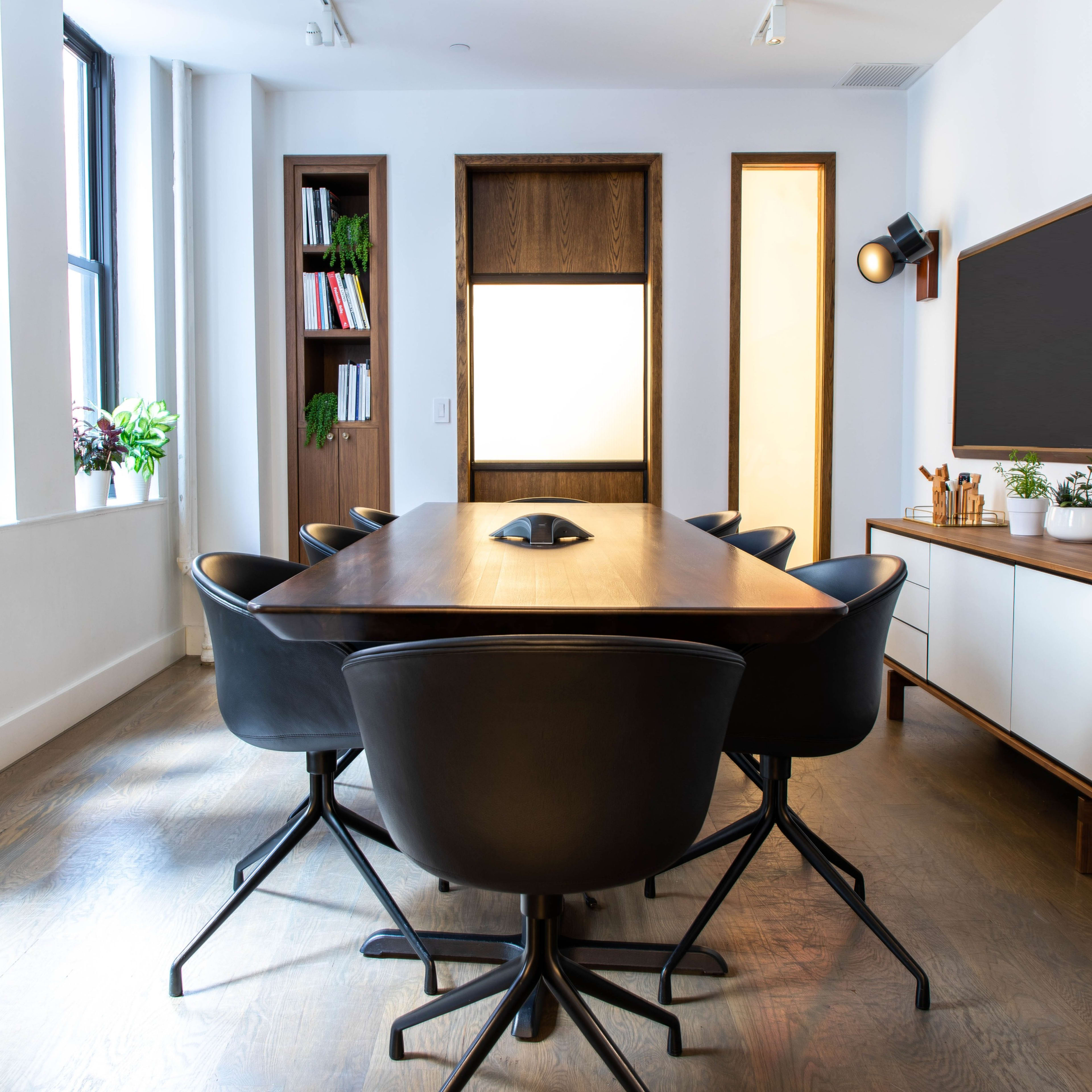 A modern conference room features a long wooden table surrounded by black swivel chairs, with large windows and built-in shelves displaying plants and books.