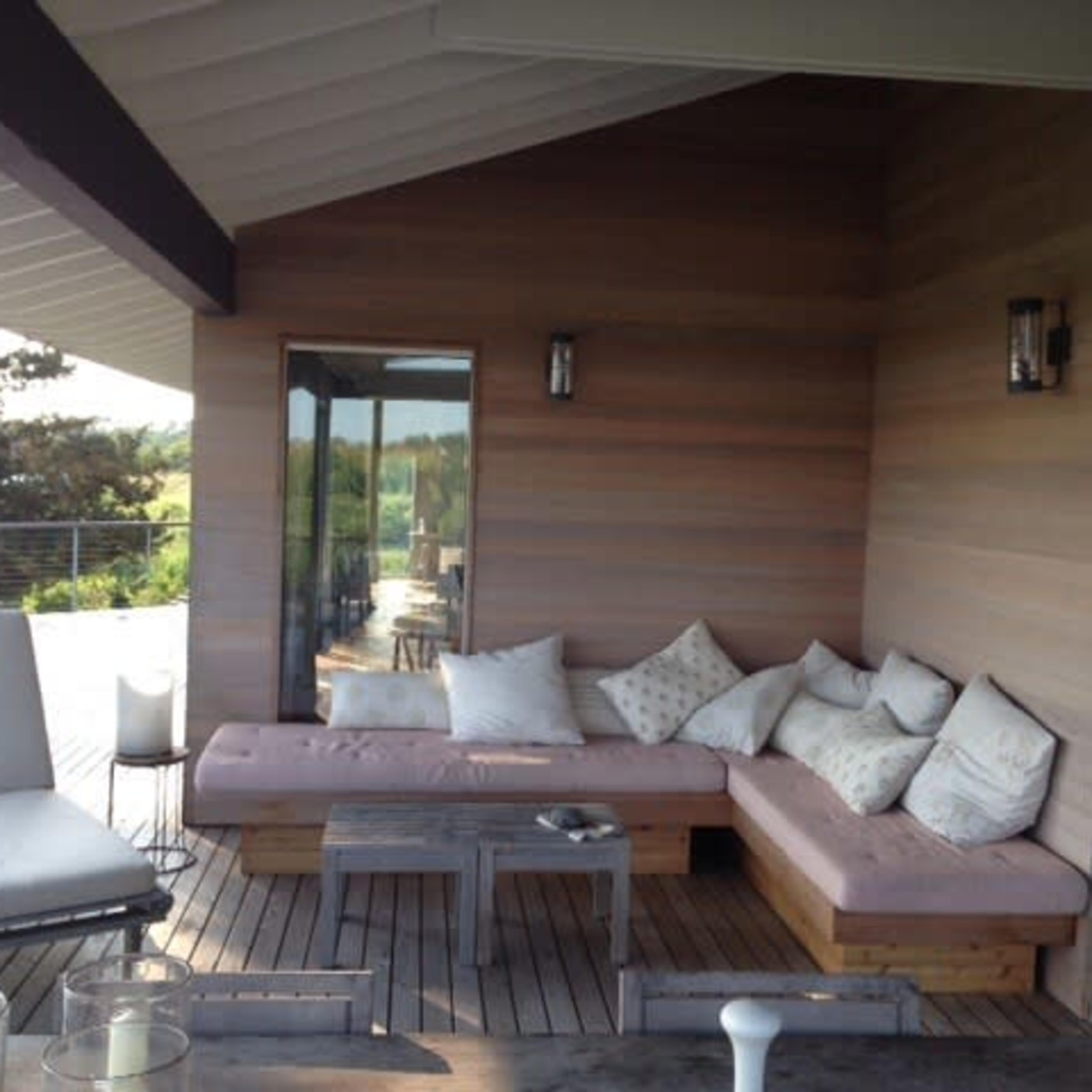 A modern outdoor seating area with a wooden deck, featuring a large cushioned sofa, a small table, and a dining space visible in the background.