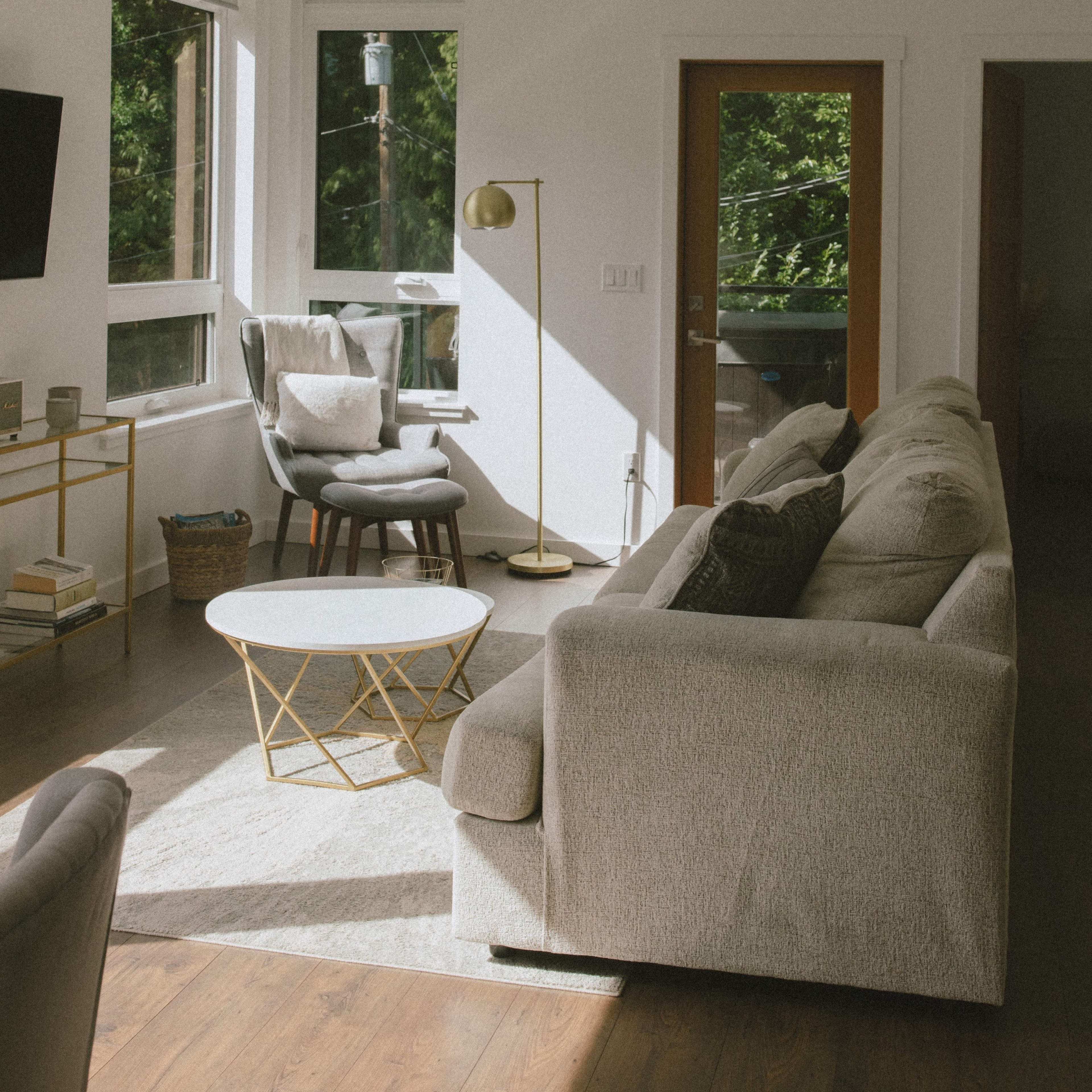 The scene shows a modern living room with large windows, a gray sofa, a round coffee table, and a floor lamp beside an armchair, leading to a door that opens to the outside.